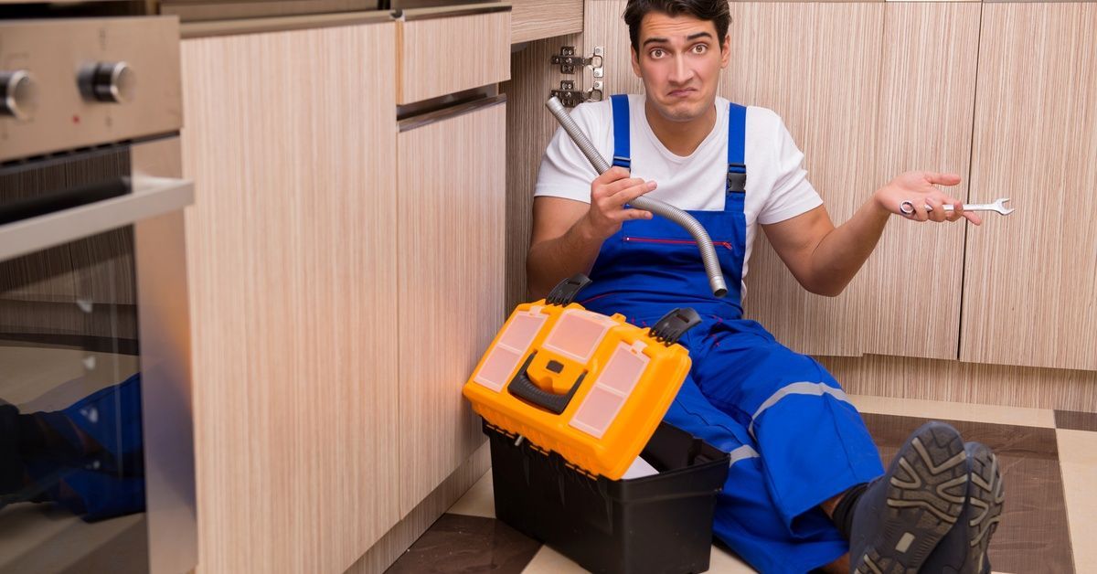A young man sitting on the floor shrugs while holding plumbing tools in both of his hands. He has his legs crossed.
