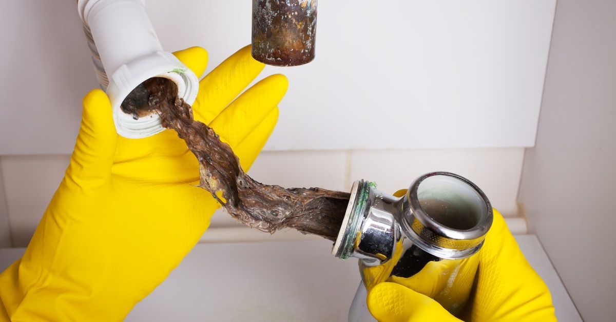 A pair of hands wearing yellow rubber gloves holds two ends of a drain pipe, revealing hair and debris.