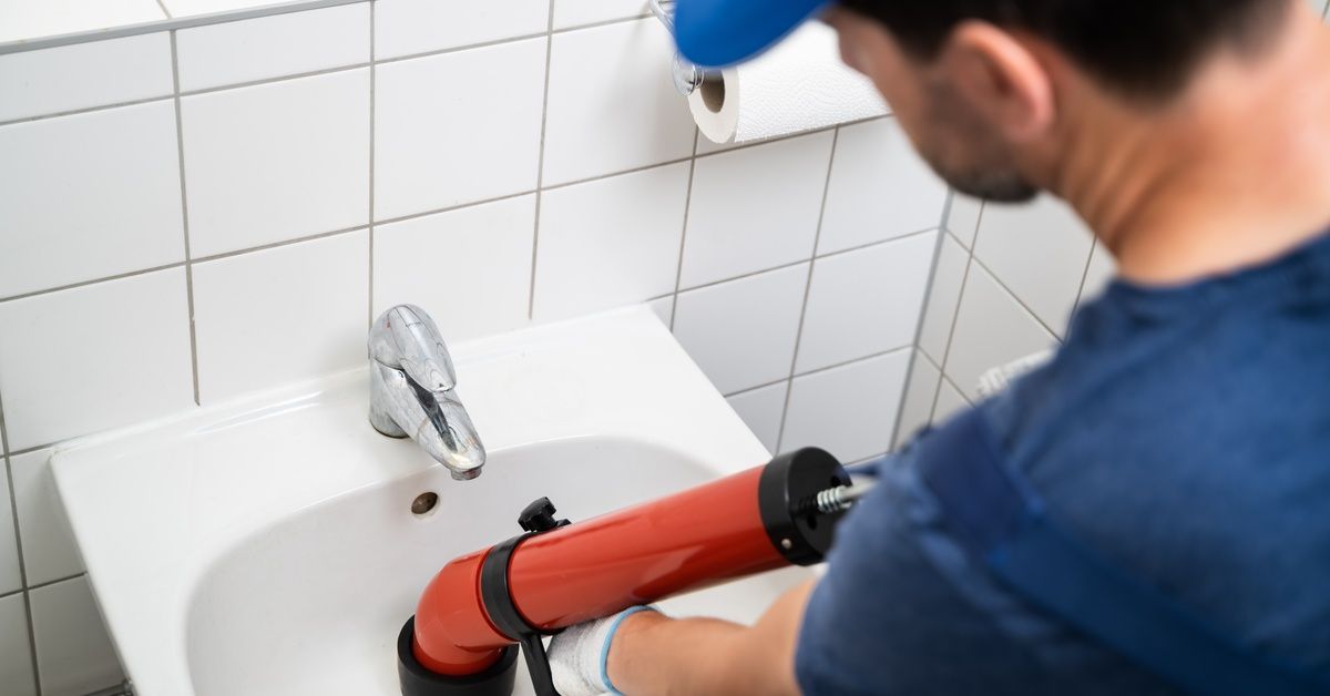 A person in a blue T-shirt and baseball cap uses a red drain tool on a sink in a bathroom with white tile.