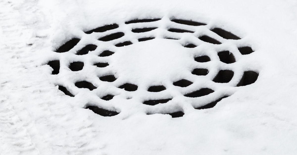 A close-up of a round sewer manhole covered in a light layer of white snow. There are not footprints on the snow.