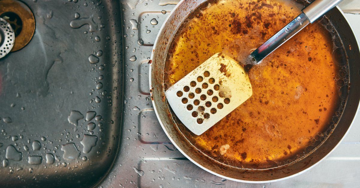 A dirty pan full of grease with a spatula resting inside it sits next to a kitchen sink with a rusty drain opening.