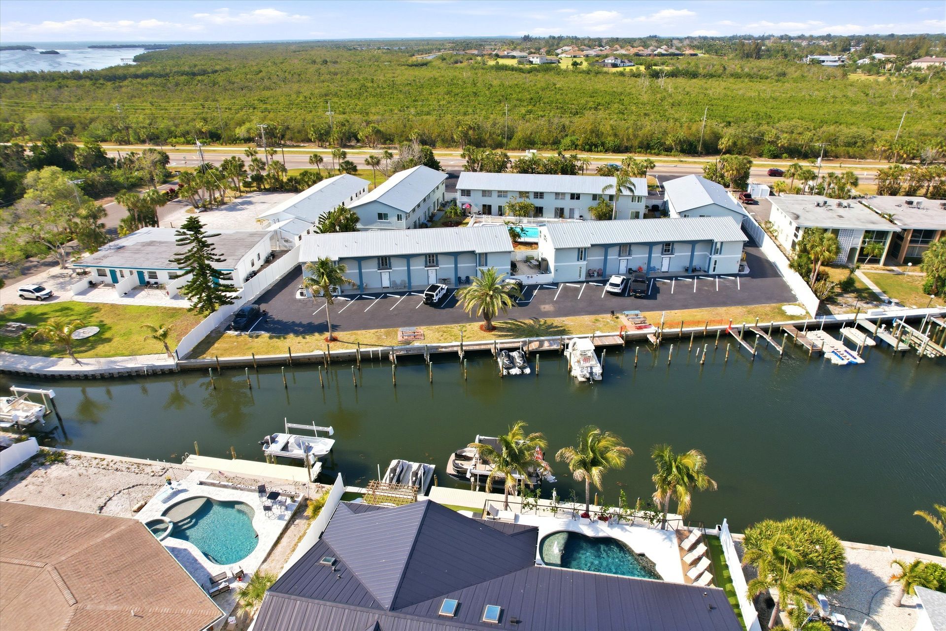 Aerial view of a waterfront resort with white buildings, canal, docks, and palm-lined pools.