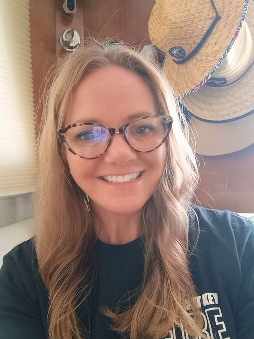 Smiling woman with glasses in a black shirt, standing indoors near a straw hat on the wall.