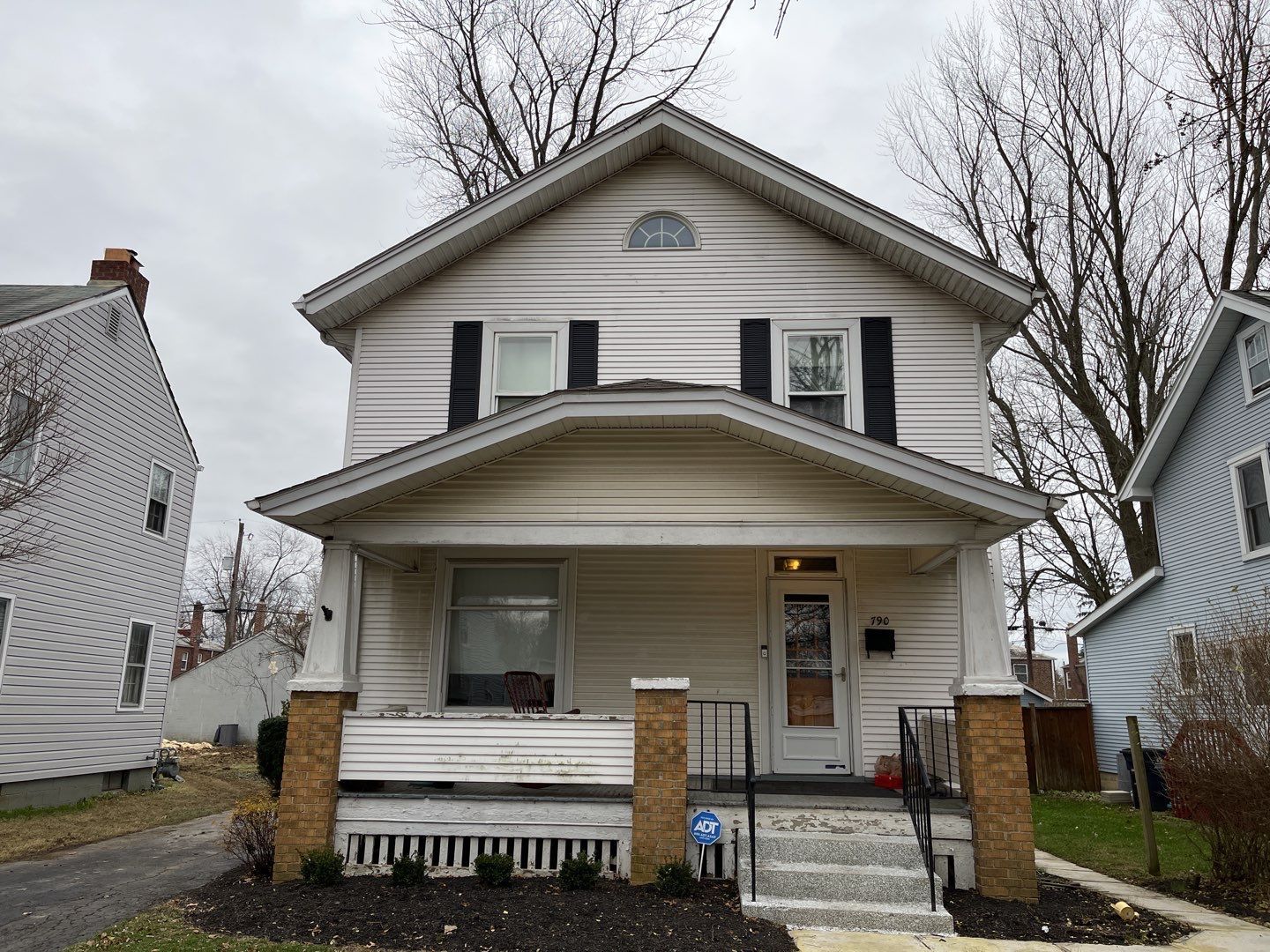 Two-story white house with a front porch and steps, framed by neighboring homes on a cloudy day