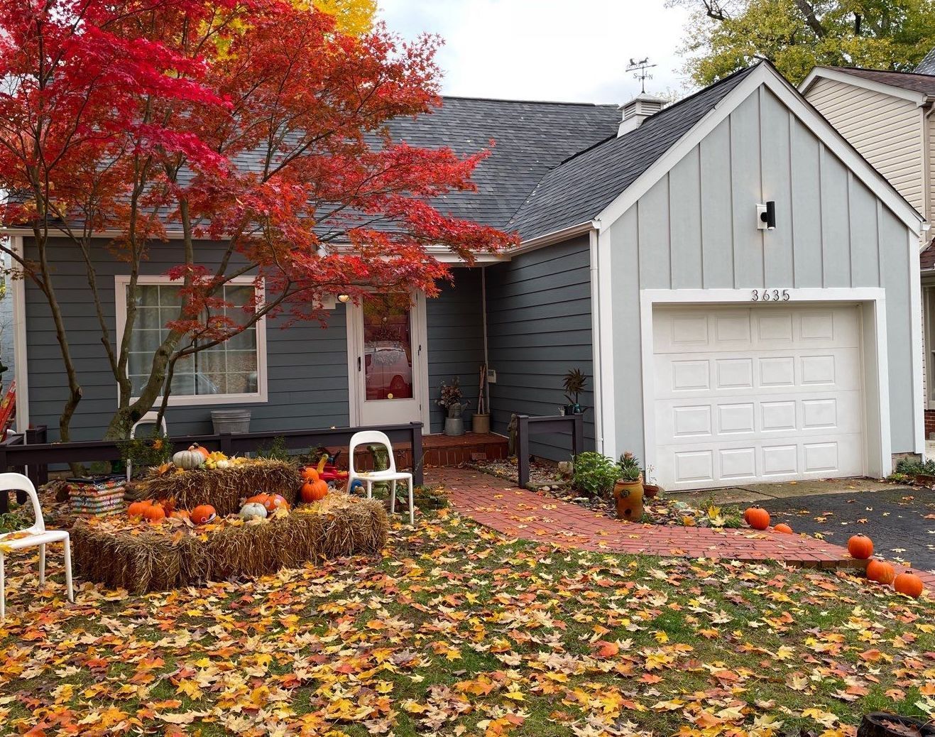 Autumn front yard with red tree, fallen leaves, gray house, and white garage door