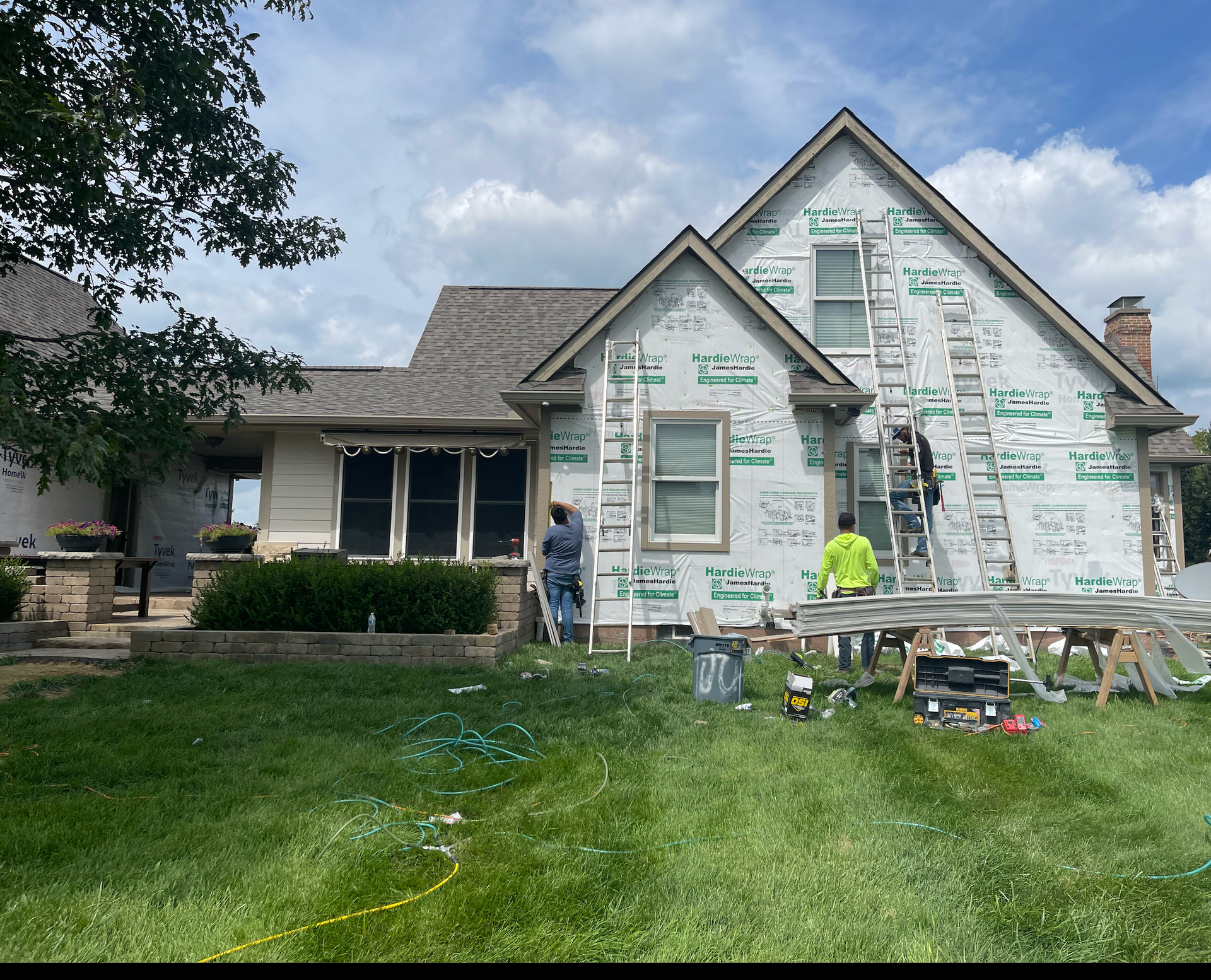 House under renovation with wrapped siding, worker in neon shirt, and tools on a front lawn
