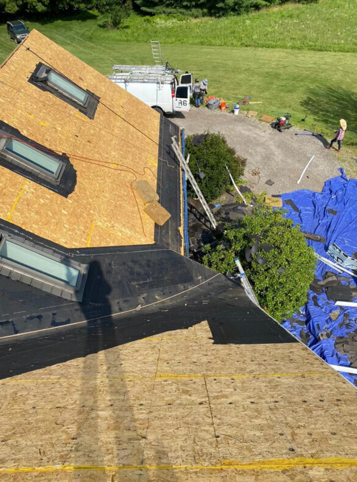 Roof under repair with workers, yellow shingles, black waterproofing, and blue tarp beside a grassy area