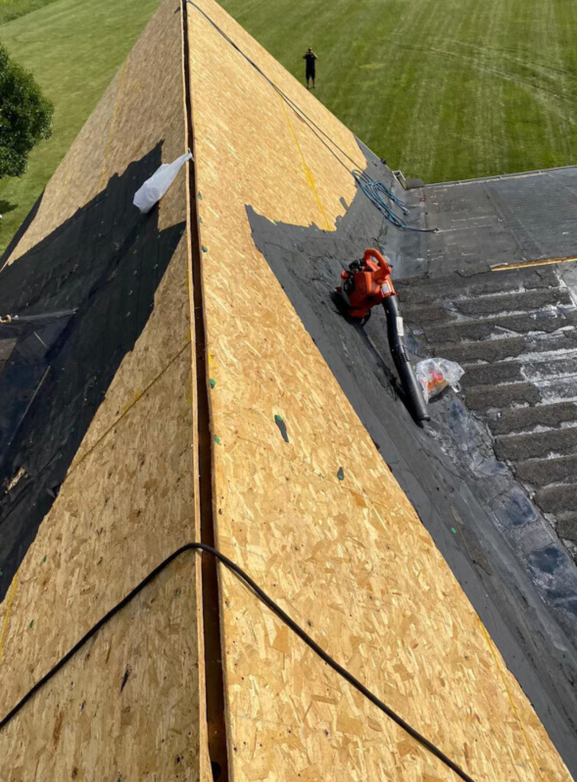Roof under repair with a worker on plywood sheathing beside the ridge line