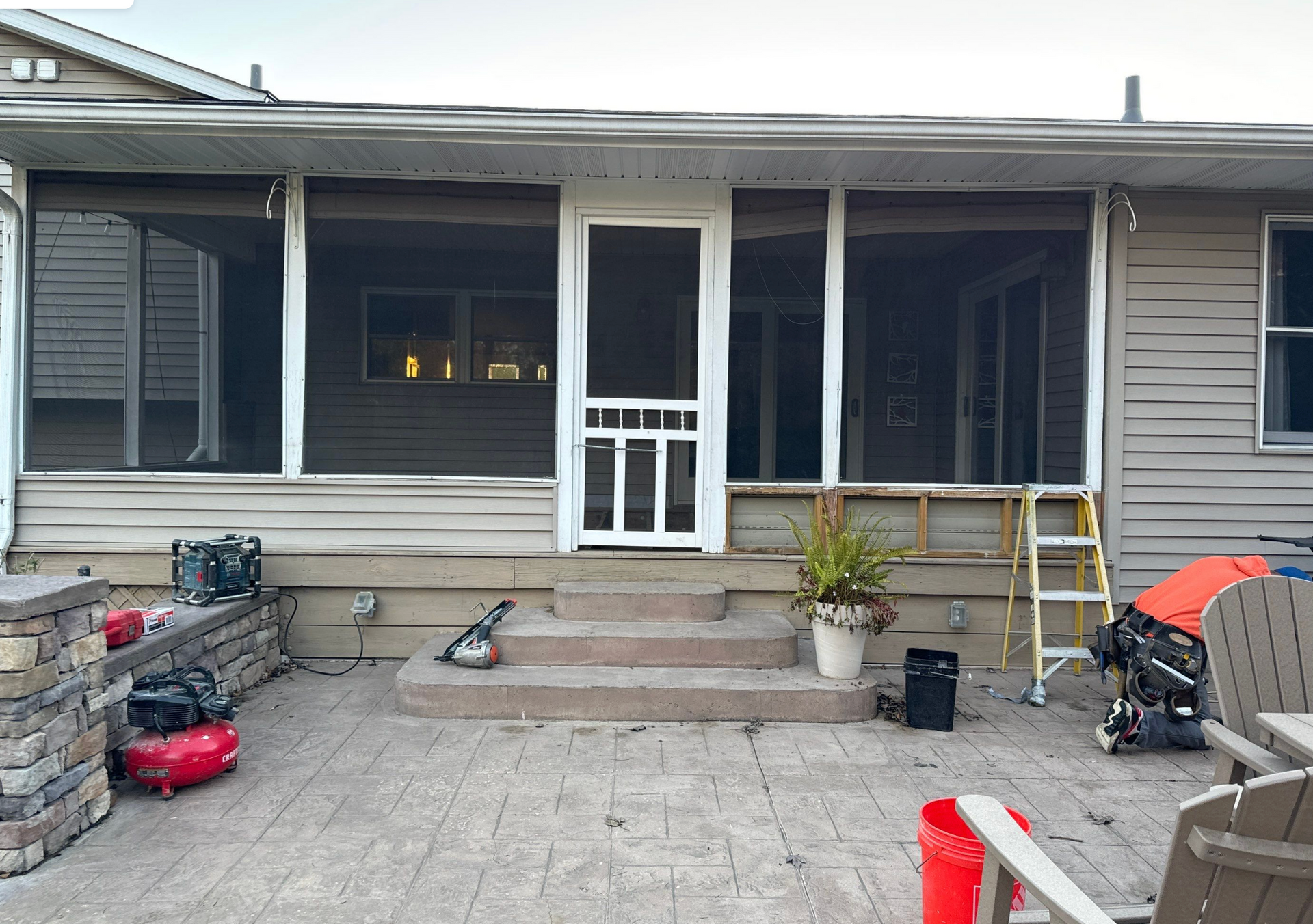 Covered backyard patio with sliding glass doors, steps, potted plant, ladder, and scattered tools on a stone patio