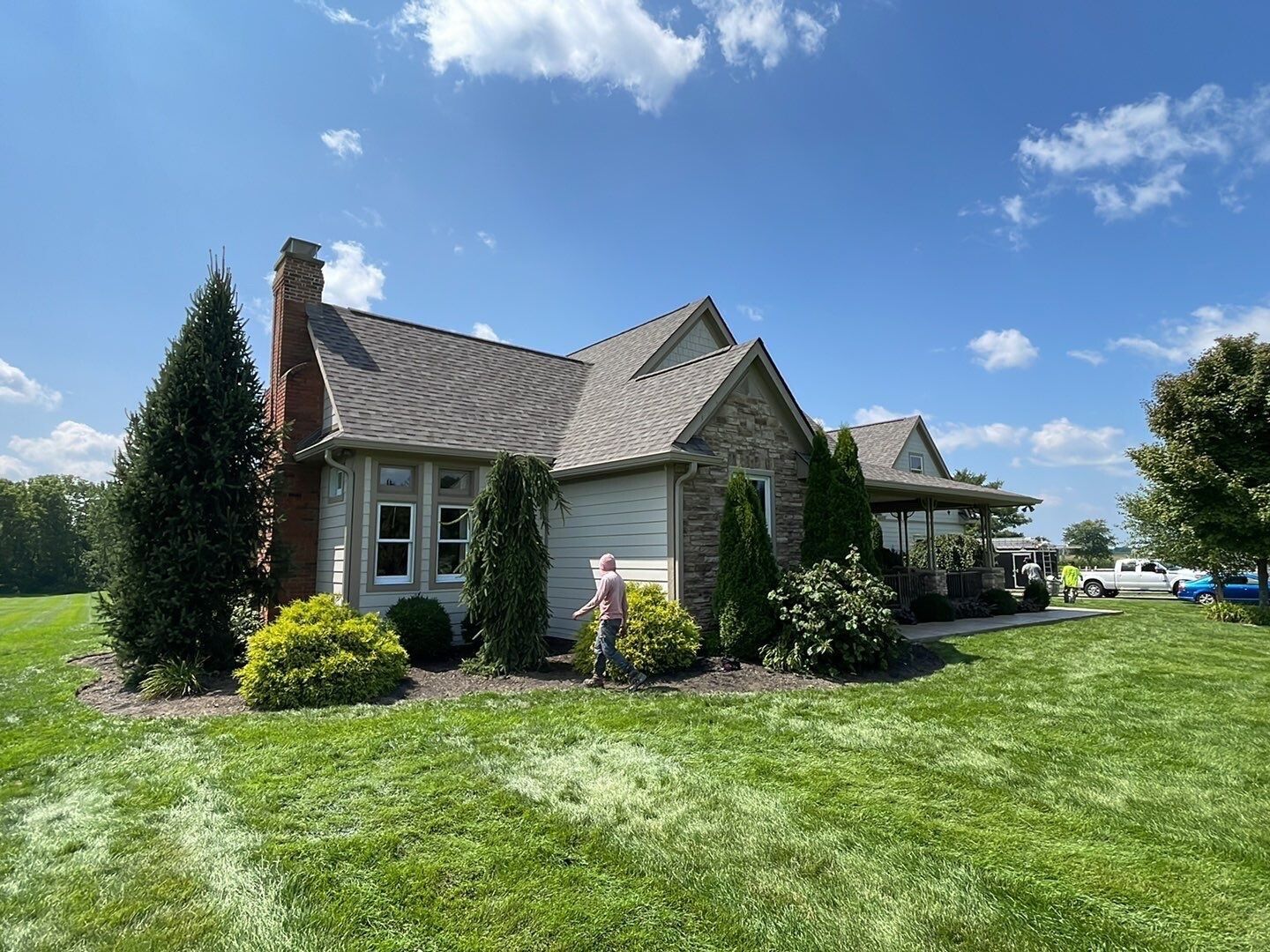 Small house with gray siding and a stone chimney in a sunny green yard under a blue sky.