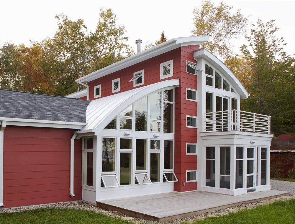 Red house with large white-framed windows, curved roofline, and a side garage surrounded by trees.