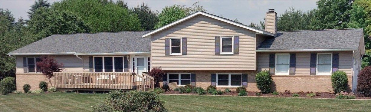 Single-story beige house with a front porch, brick foundation, and a chimney, set behind a lawn and trees.