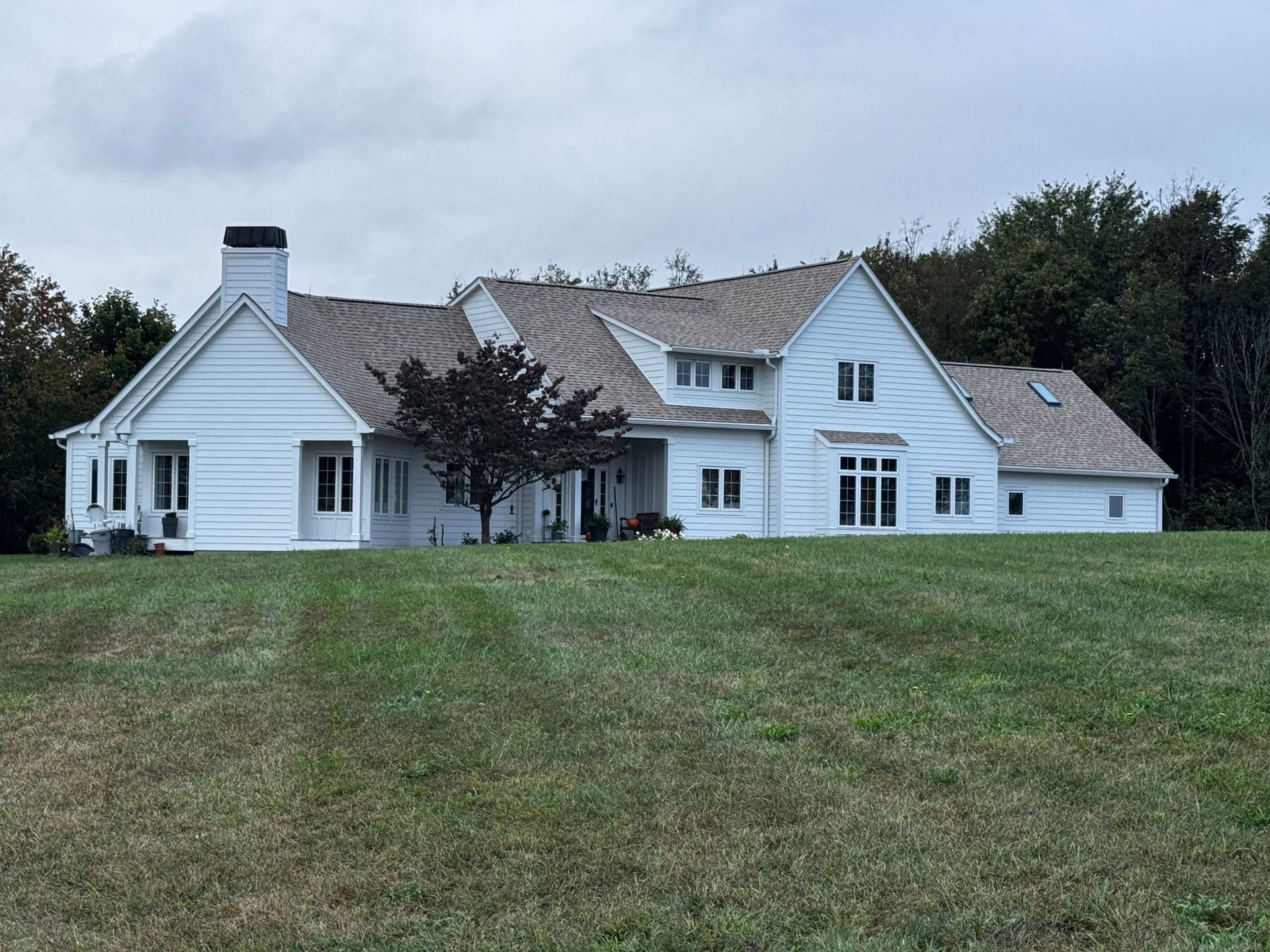 Large white farmhouse with gray roof on a grassy hill under an overcast sky