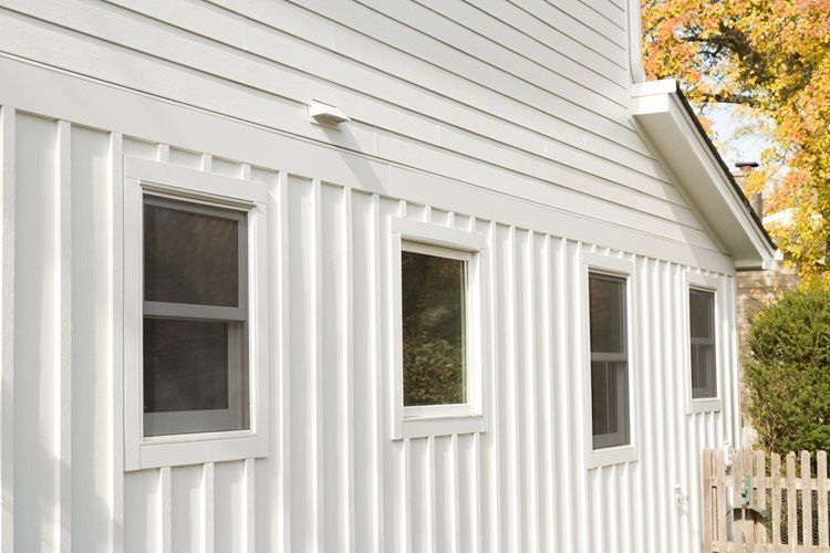 White house exterior with three windows and vertical siding, beside a small picket fence and autumn trees