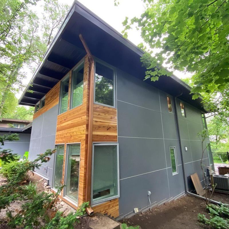 Modern two-story house with gray siding, warm wood accents, and large windows surrounded by trees