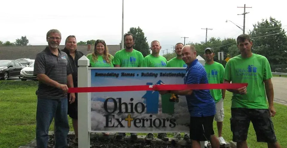 Group in green shirts at Ohio Exteriors sign, holding a red ribbon at an outdoor ribbon-cutting event