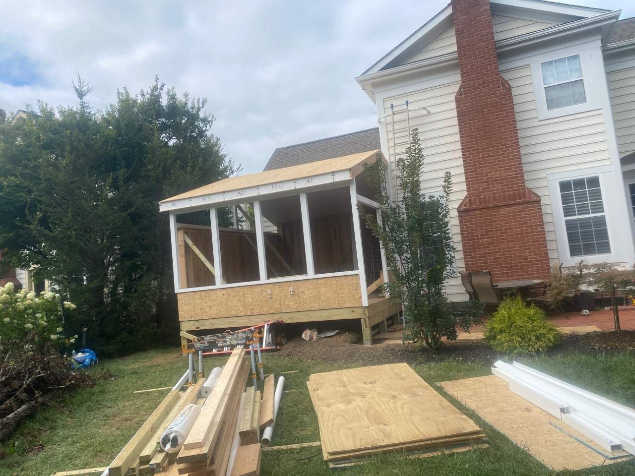 Back porch construction beside a white house, with lumber and building materials in the yard