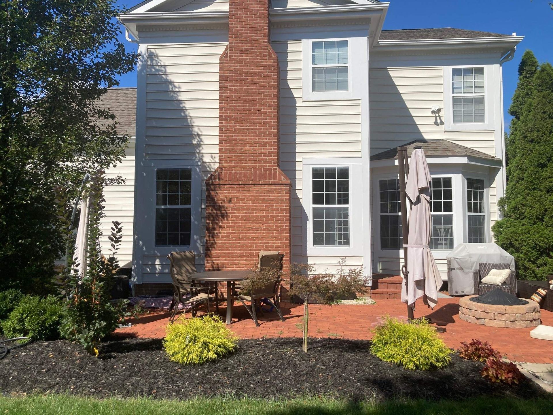 Backyard patio with white house, red brick chimney, black mulch, and potted plants under bright sun