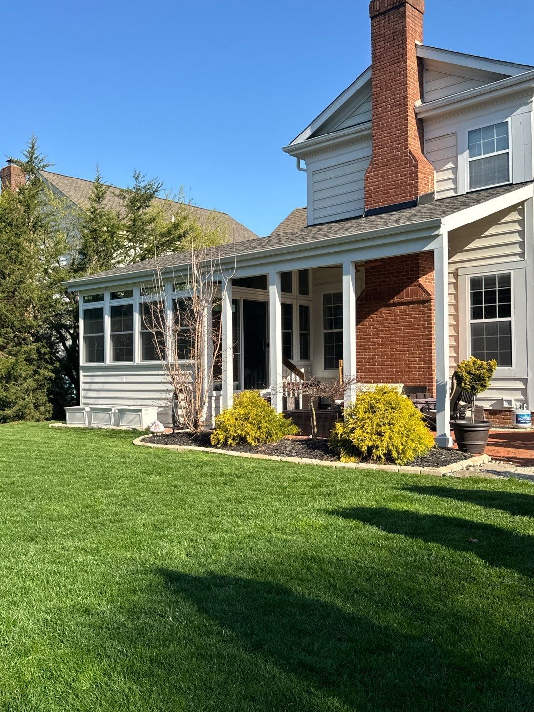 Sunlit suburban house with white porch, red brick chimney, and green lawn