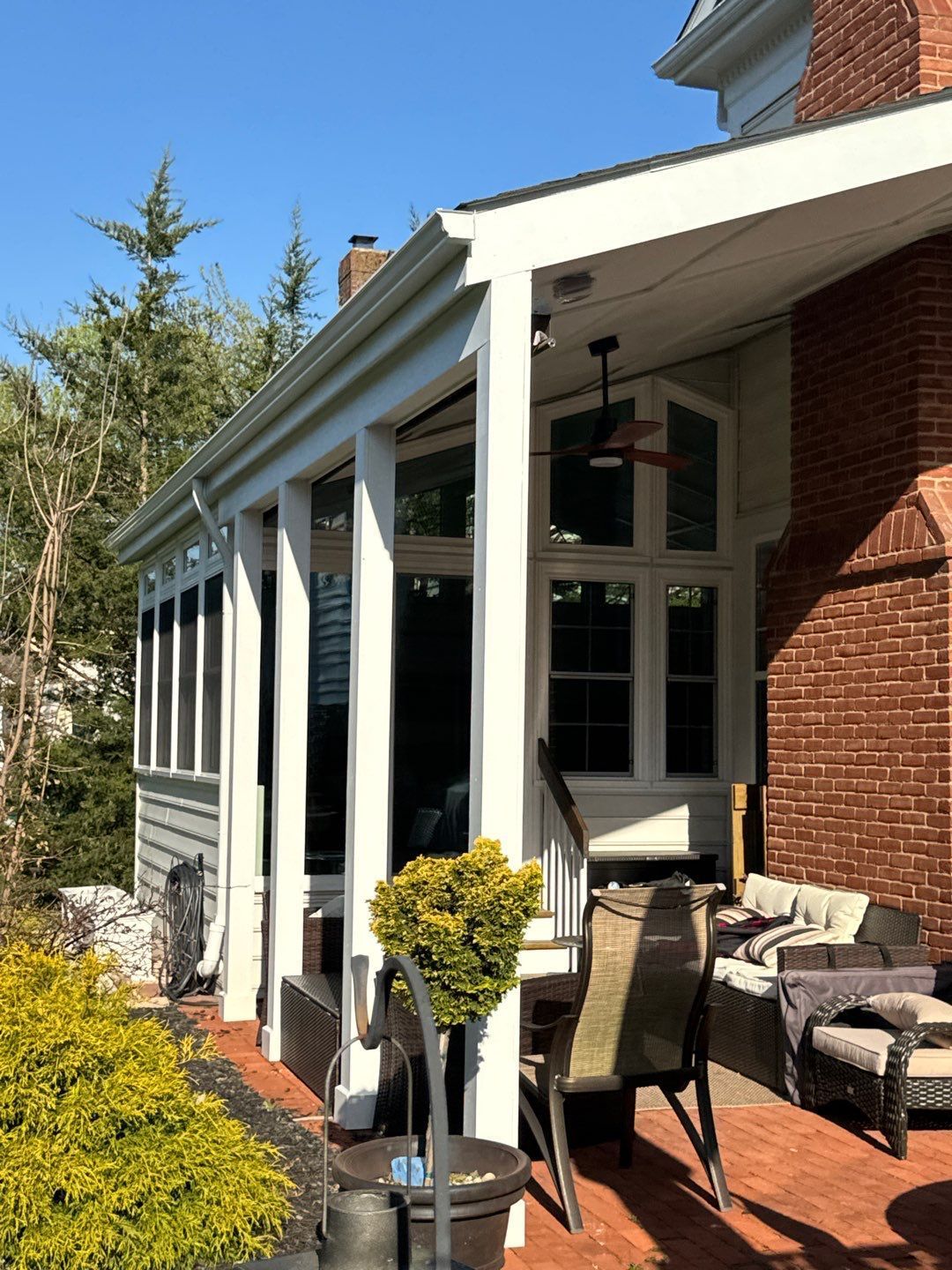 Covered patio with white columns, wicker chairs, potted plants, and a brick house exterior in sunlight