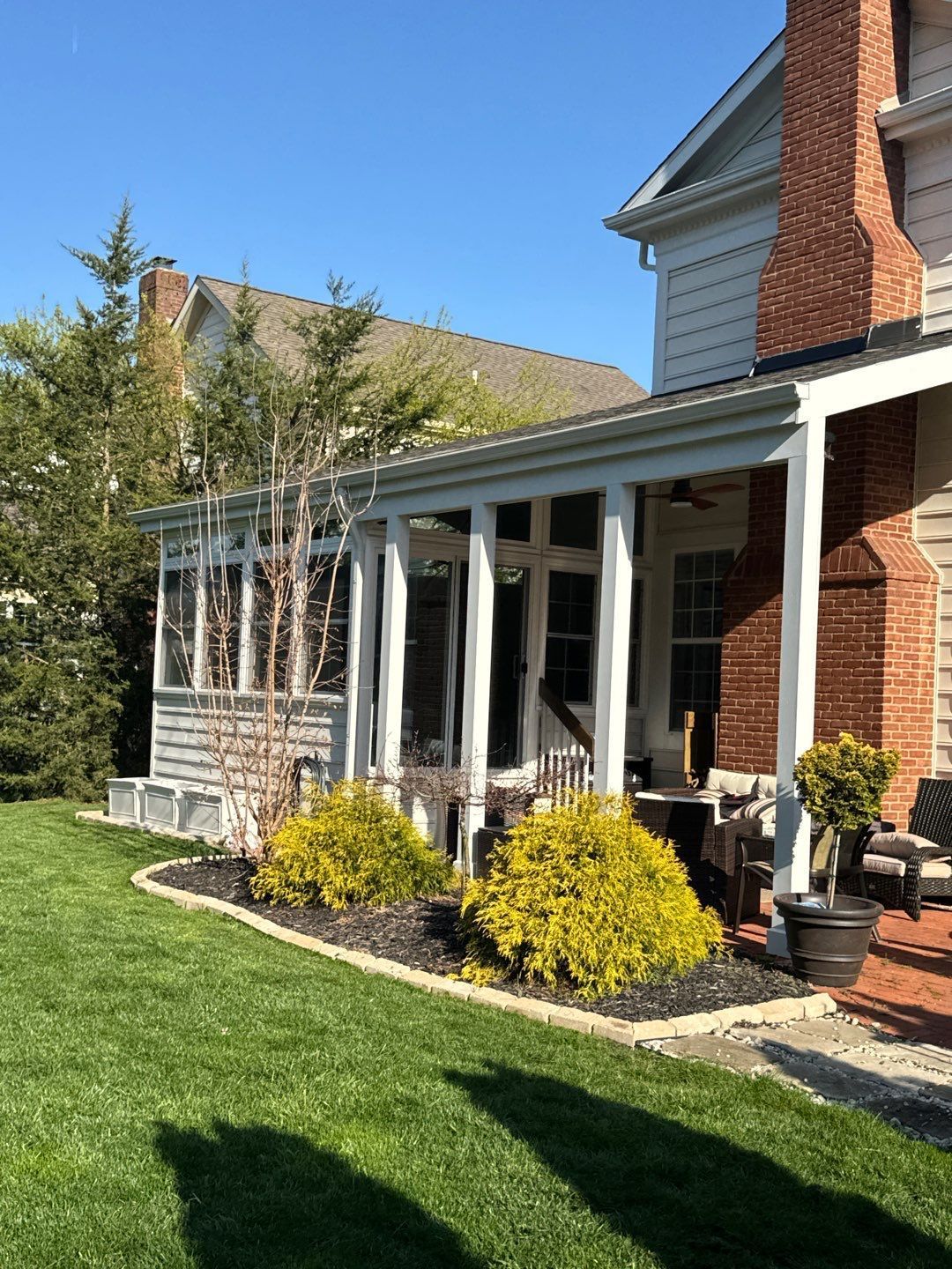 Brick house porch with white columns, yellow shrubs, and a green lawn under a blue sky