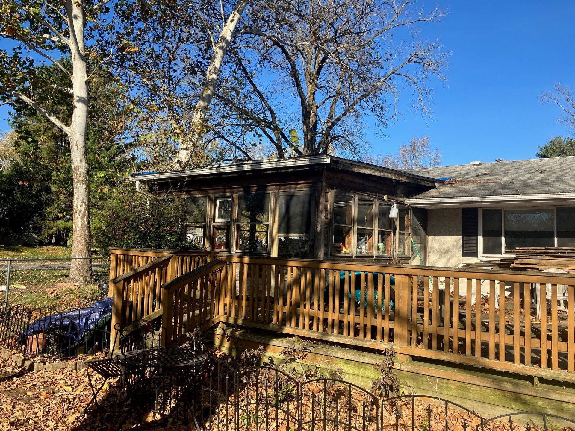 Wooden deck and railing beside a house with a screened porch, under leafless trees on a sunny day