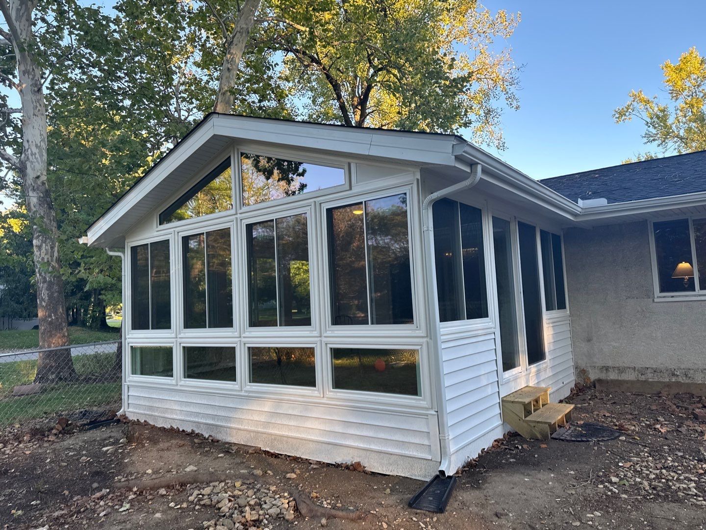Small white sunroom addition with large windows and a gabled roof beside a house in a wooded yard