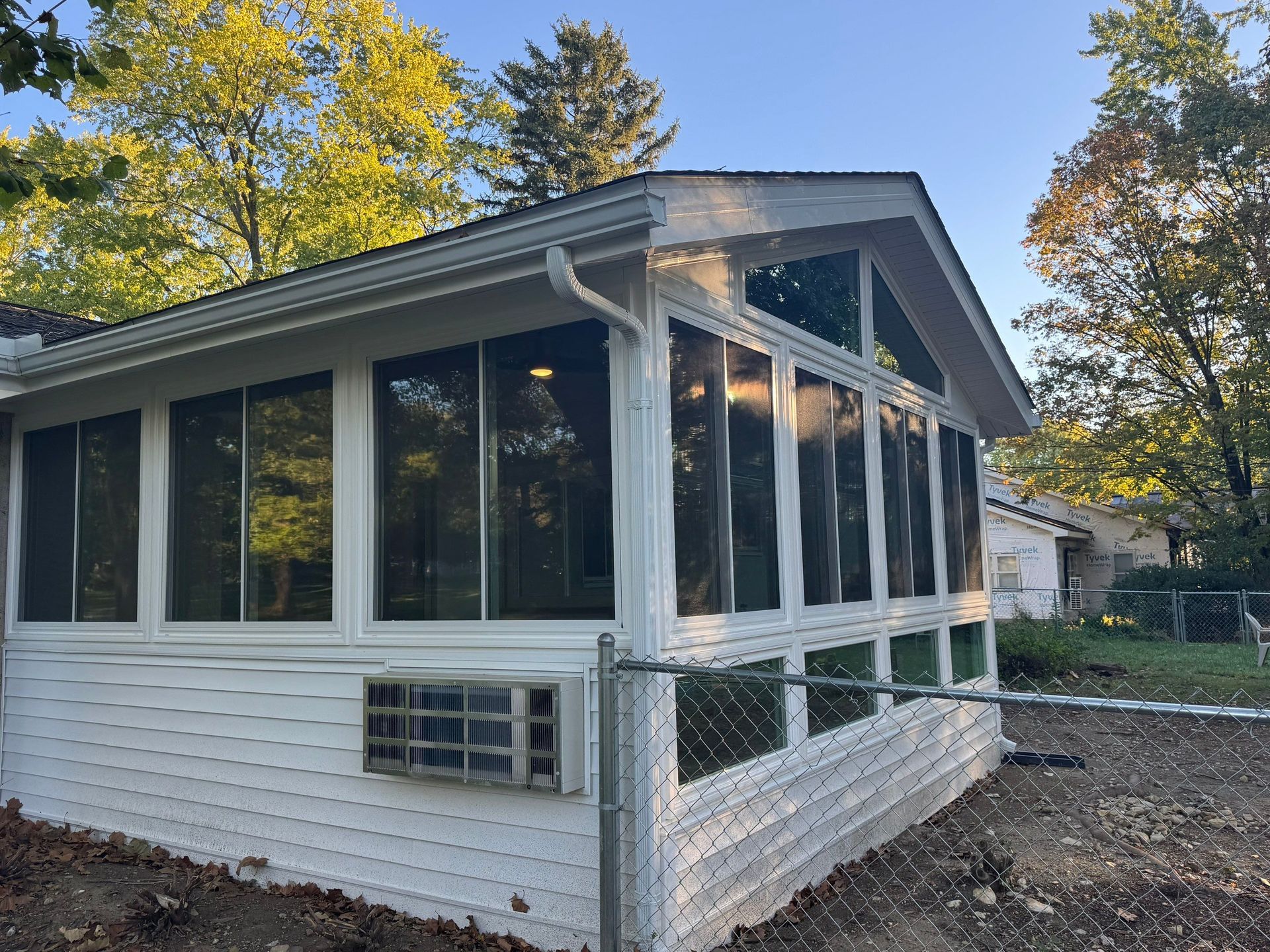 White screened porch with dark roof and windows on a house, surrounded by trees in fall.