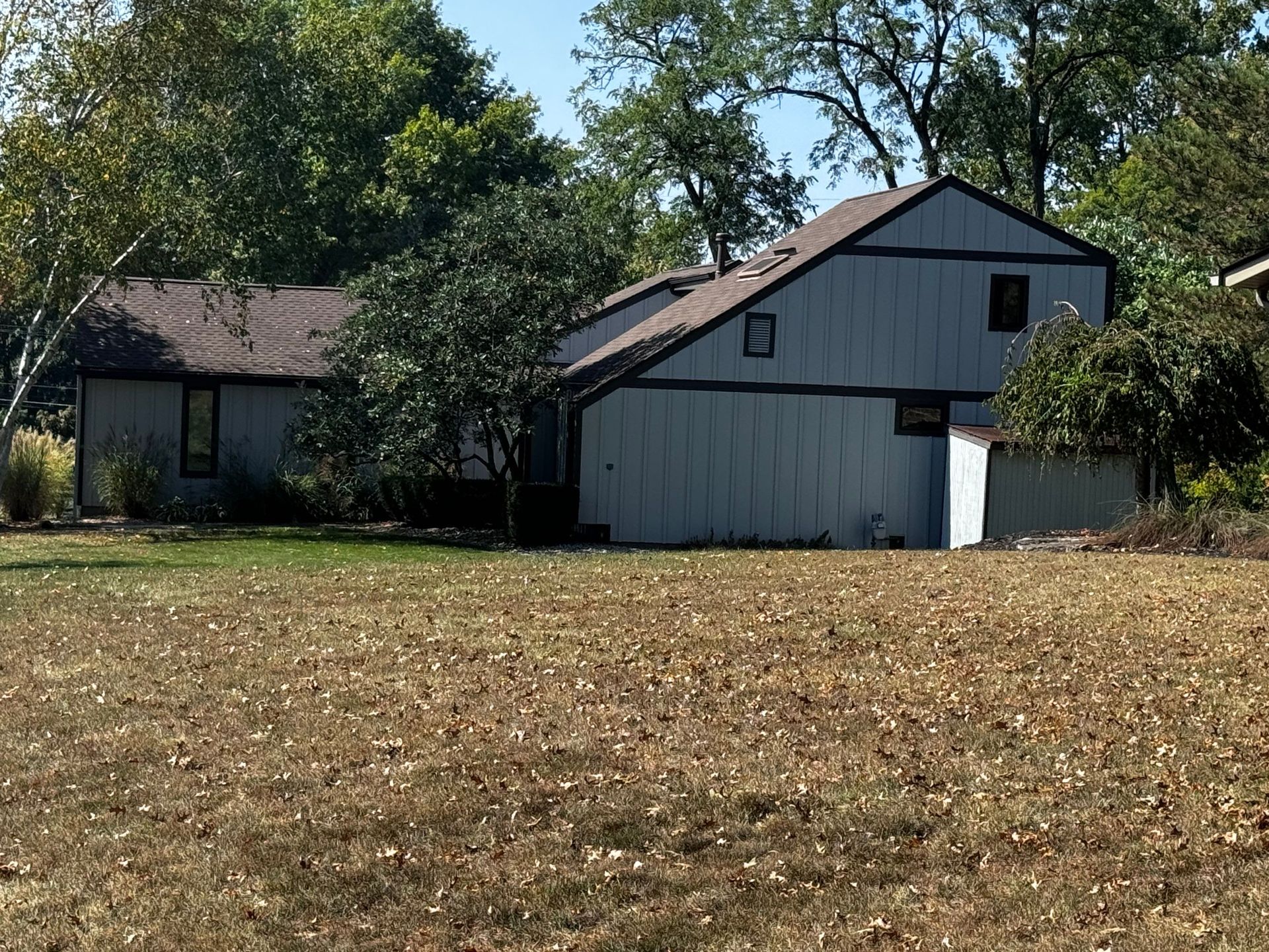 Two-story gray house with a brown roof beside a smaller building, set behind a dry grassy field and trees.