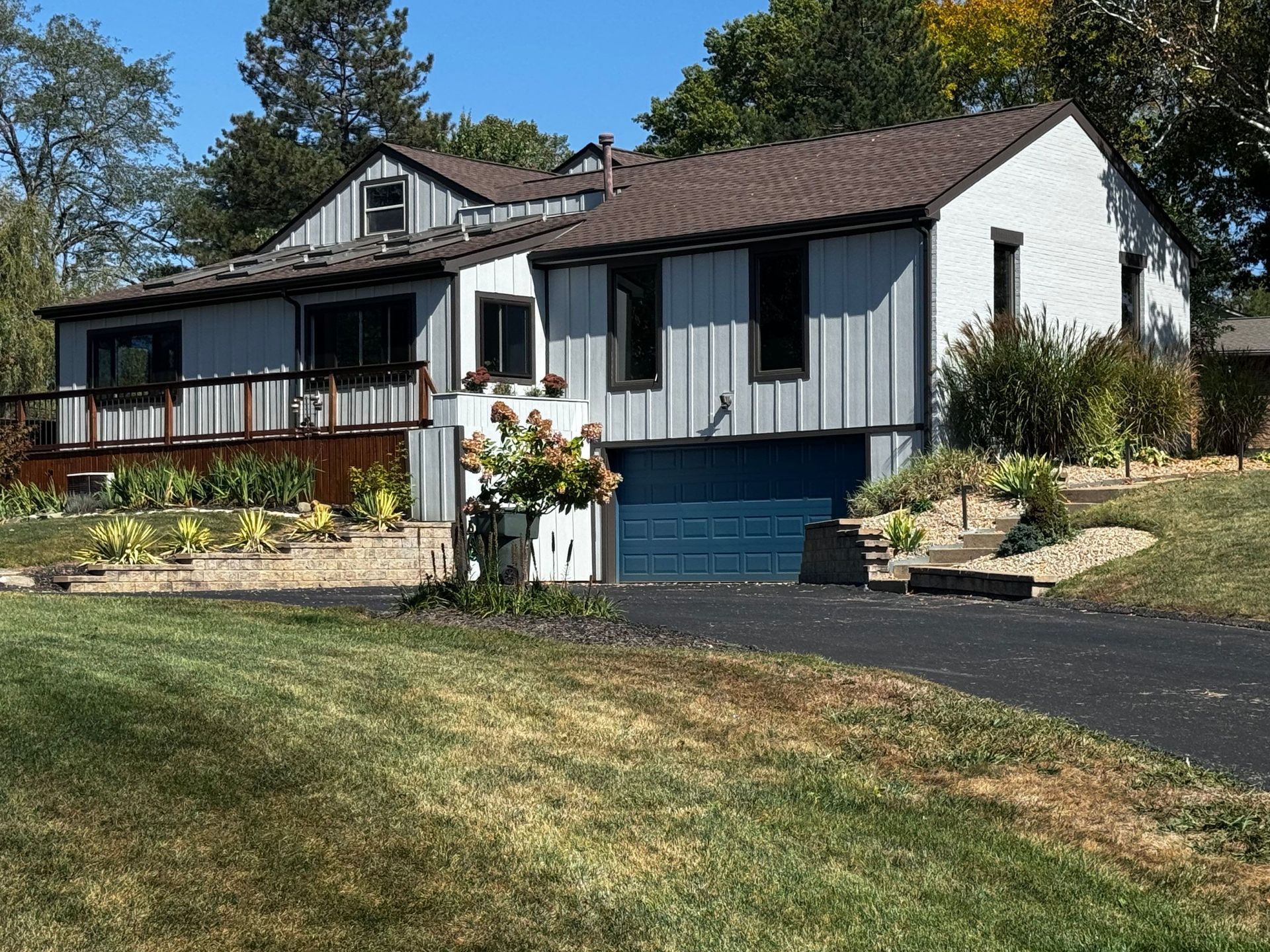 Single-story house with blue garage door, gray siding, and a front porch on a sunny hillside