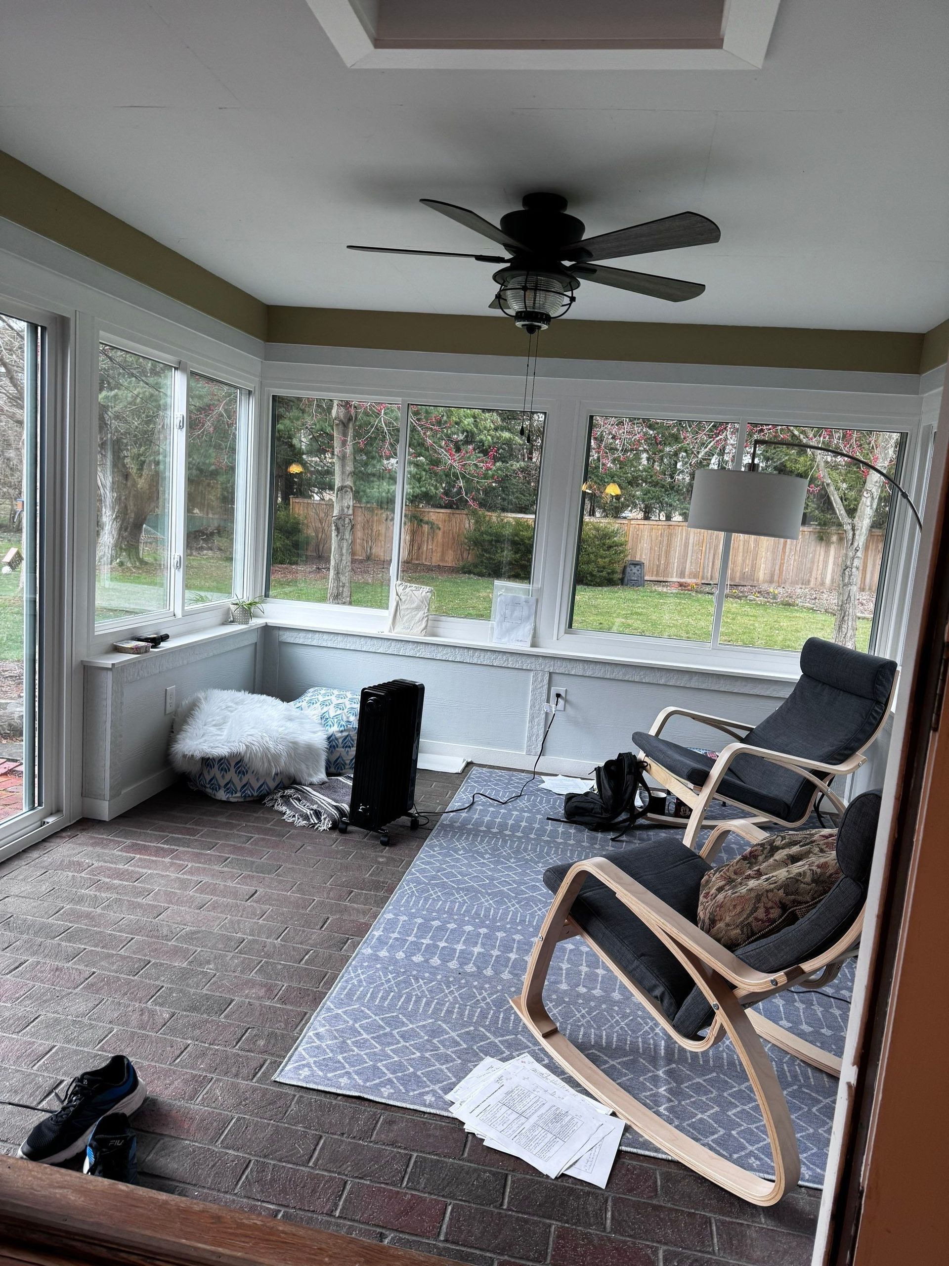 Enclosed porch with large windows, ceiling fan, gray chairs, and a blue rug; clutter and papers on the floor
