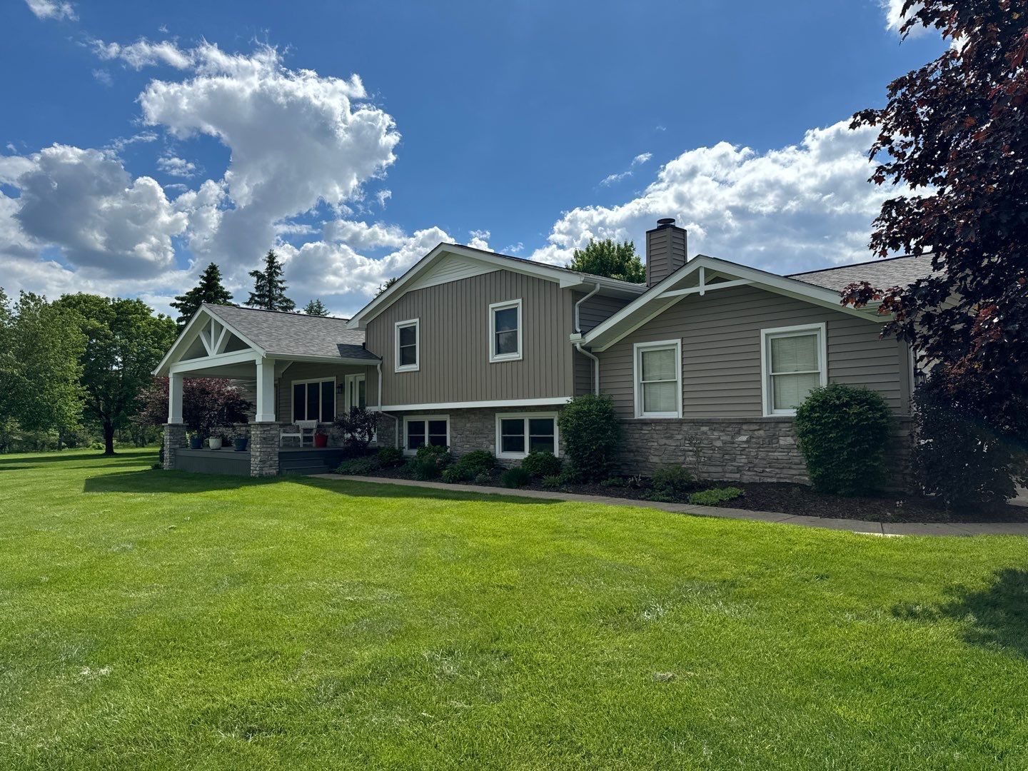 Suburban house with beige siding, covered porch, and a large green lawn under a partly cloudy sky