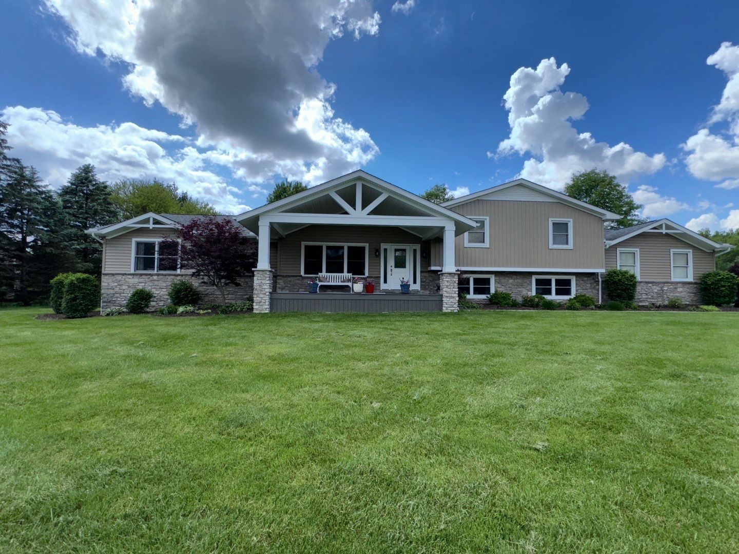 Large beige house with a covered porch and wide green lawn under a partly cloudy blue sky