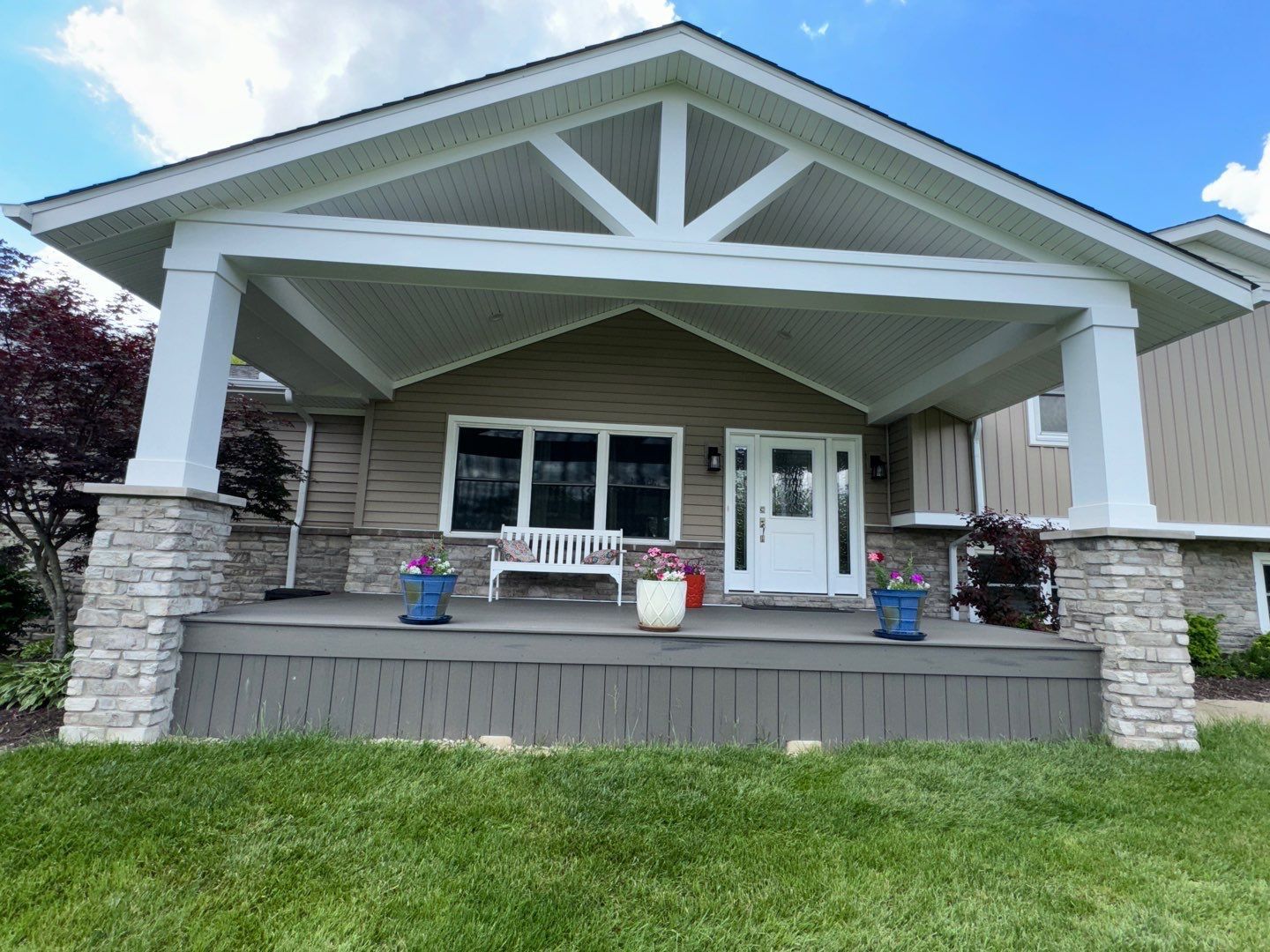 Front porch of a house with white railings, stone columns, and potted flowers under a gray roof