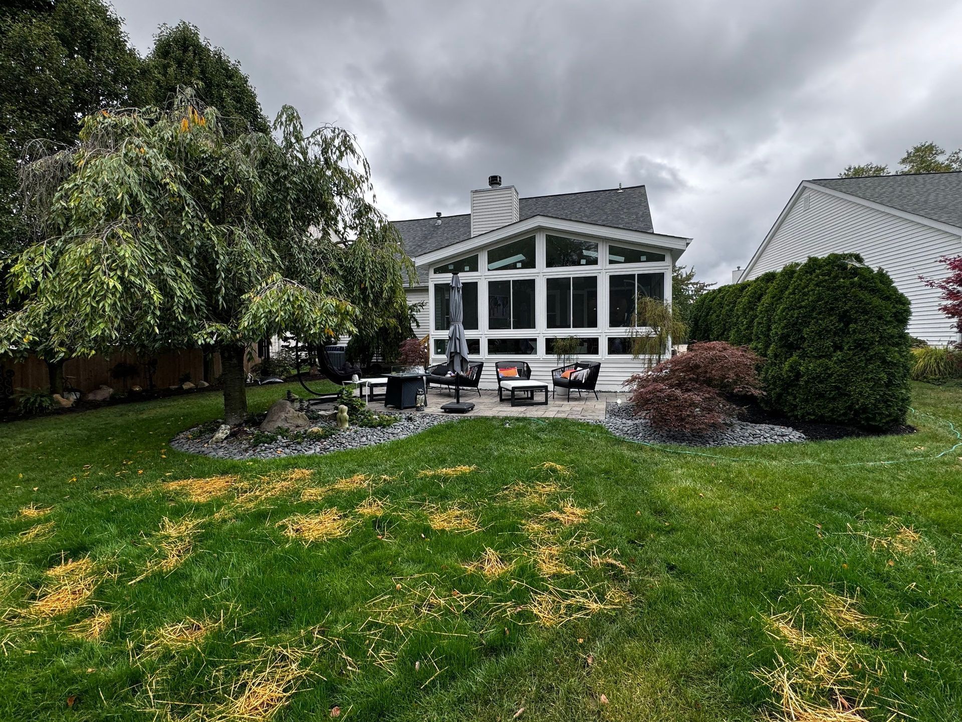 Backyard patio with glass sunroom, outdoor seating, and a large green lawn under cloudy skies