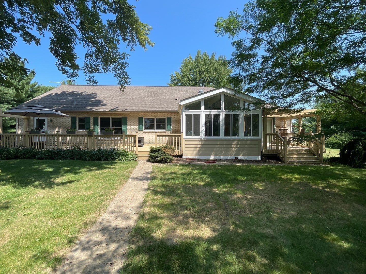Single-story beige house with a glass sunroom, front porch, and shaded lawn under blue sky