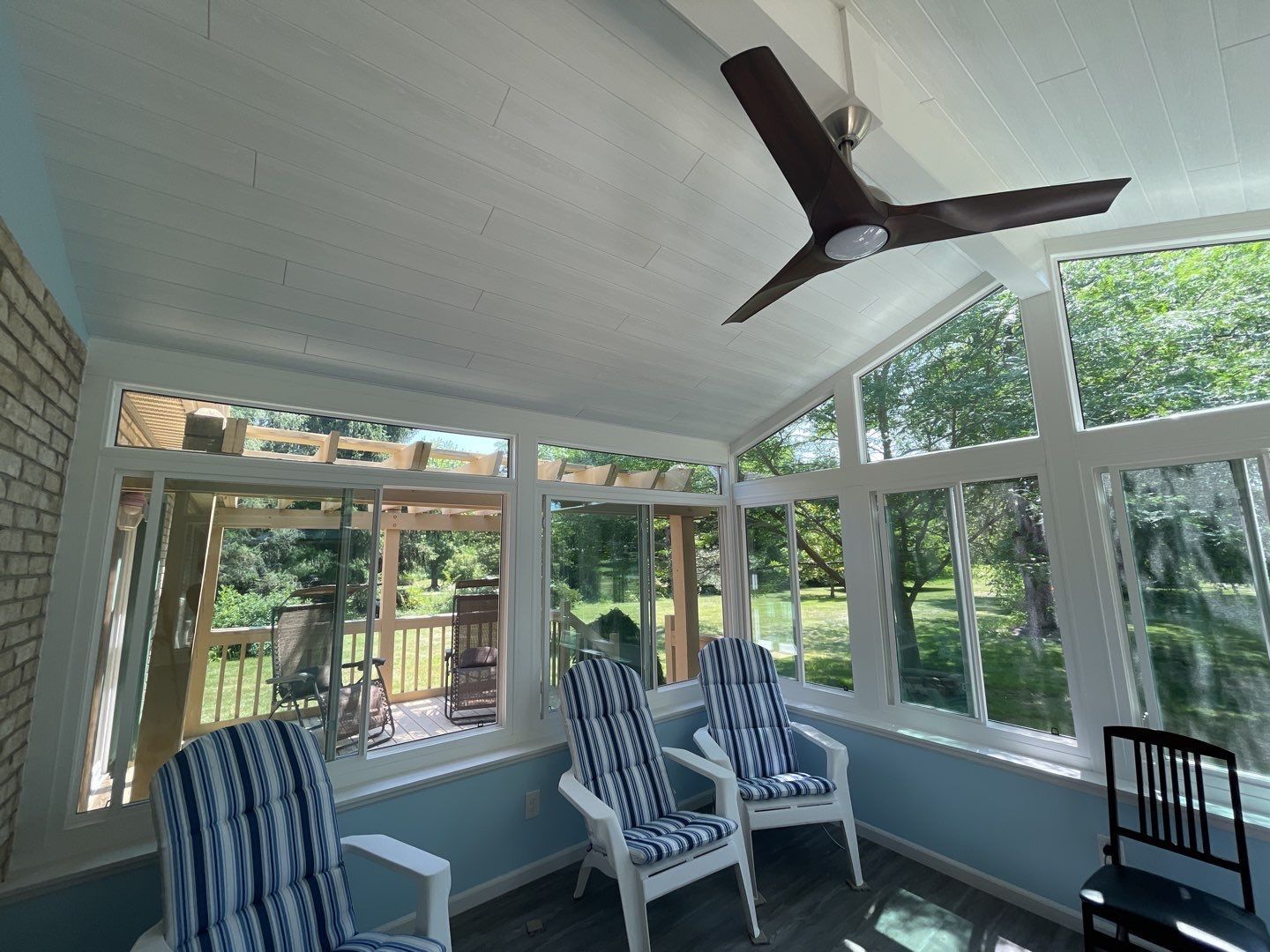 Sunroom with white ceiling, dark ceiling fan, blue chairs, and large windows overlooking greenery
