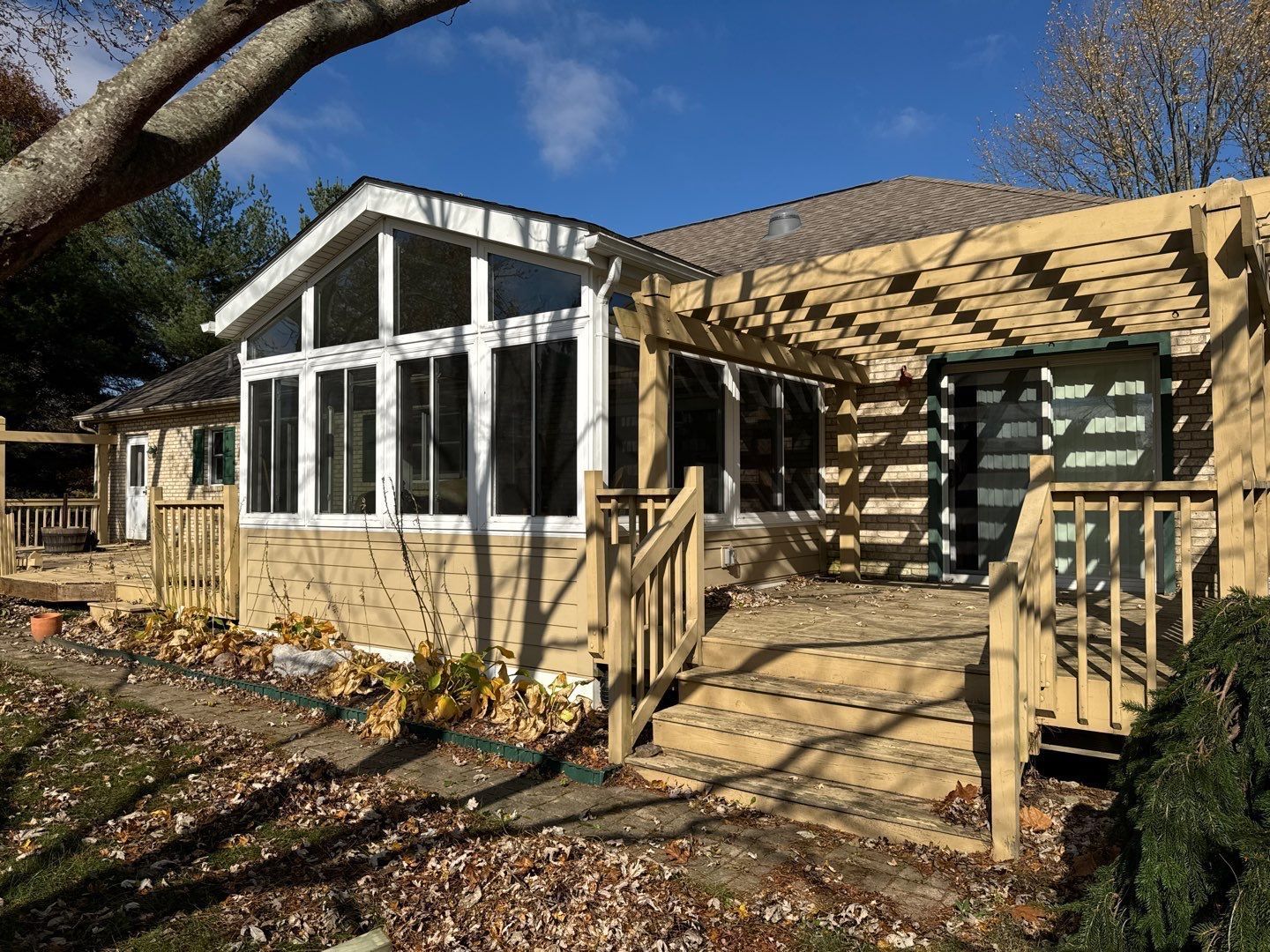 Stone house with a sunroom, wooden deck railings, and a pergola under a clear blue sky.
