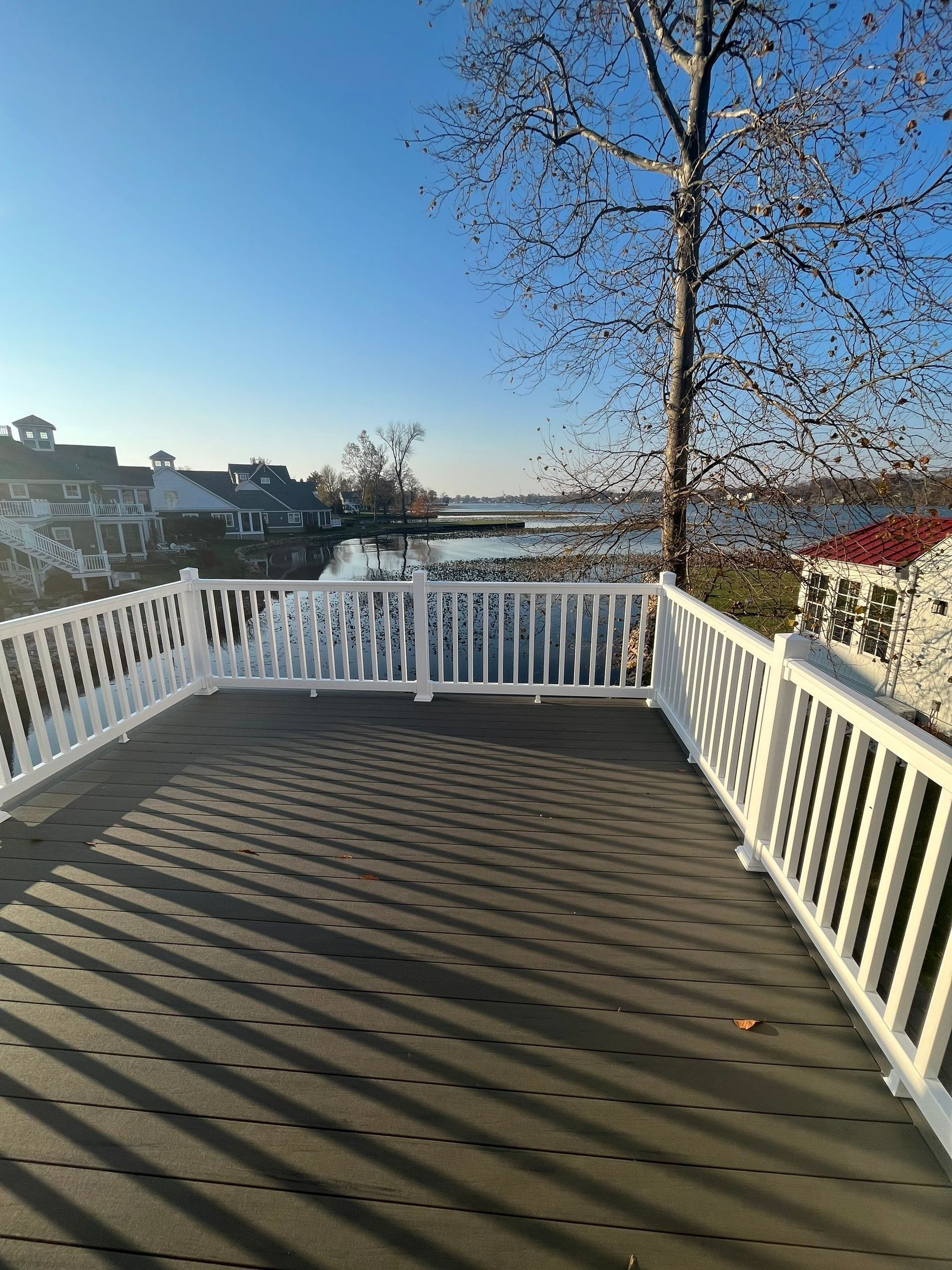 Wooden deck with white railing overlooking a river under a clear blue sky, long shadows across the boards