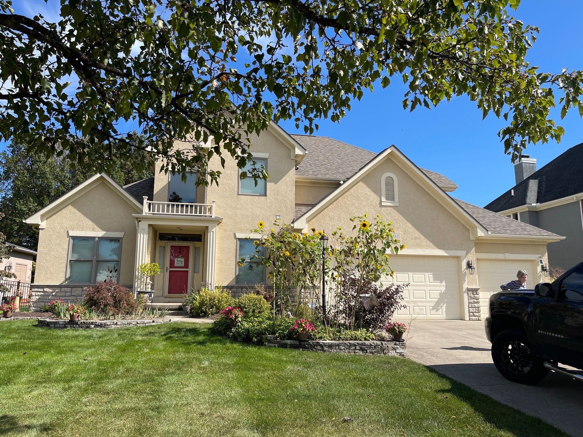 Suburban two-story beige house with red front door, driveway, lawn, and parked black SUV under a tree