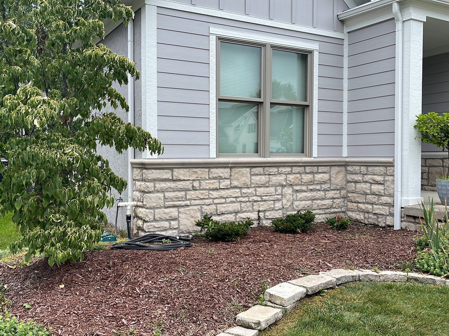 Gray house exterior with stone foundation, window, and landscaped mulch bed with shrubs and edging stones