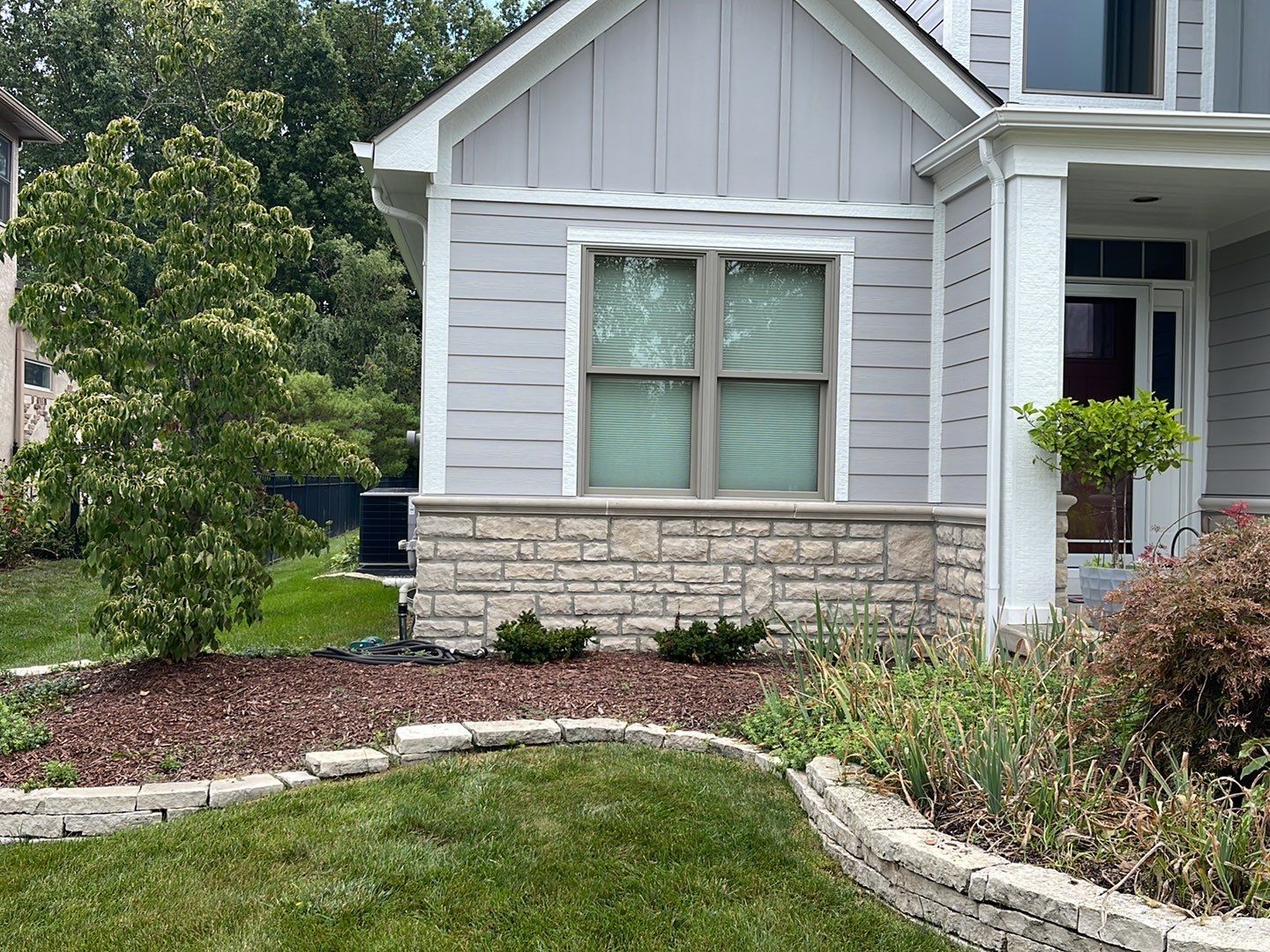 Front of a gray house with stone trim, a window, and landscaped flower beds by a curved lawn edge