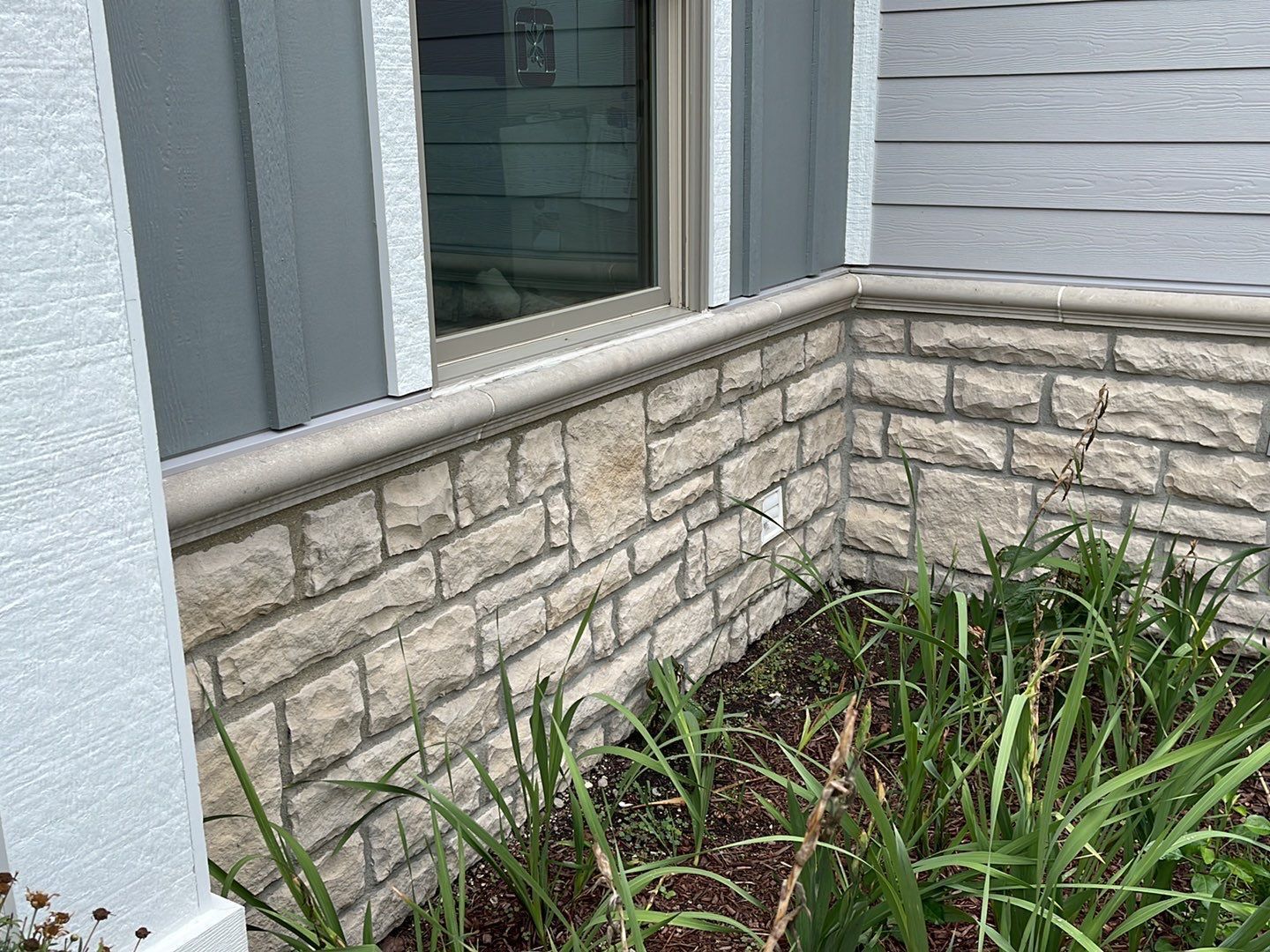 Stone exterior wall beside a window, with green plants in a small garden bed.