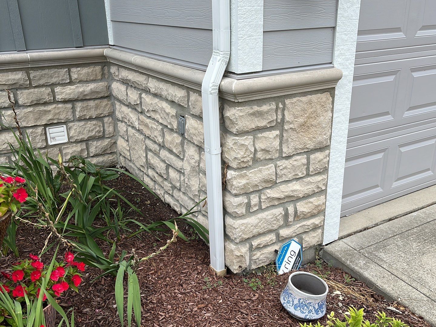 Stone house corner beside garage door, with downspout, flowers, and a small birdbath in mulch.