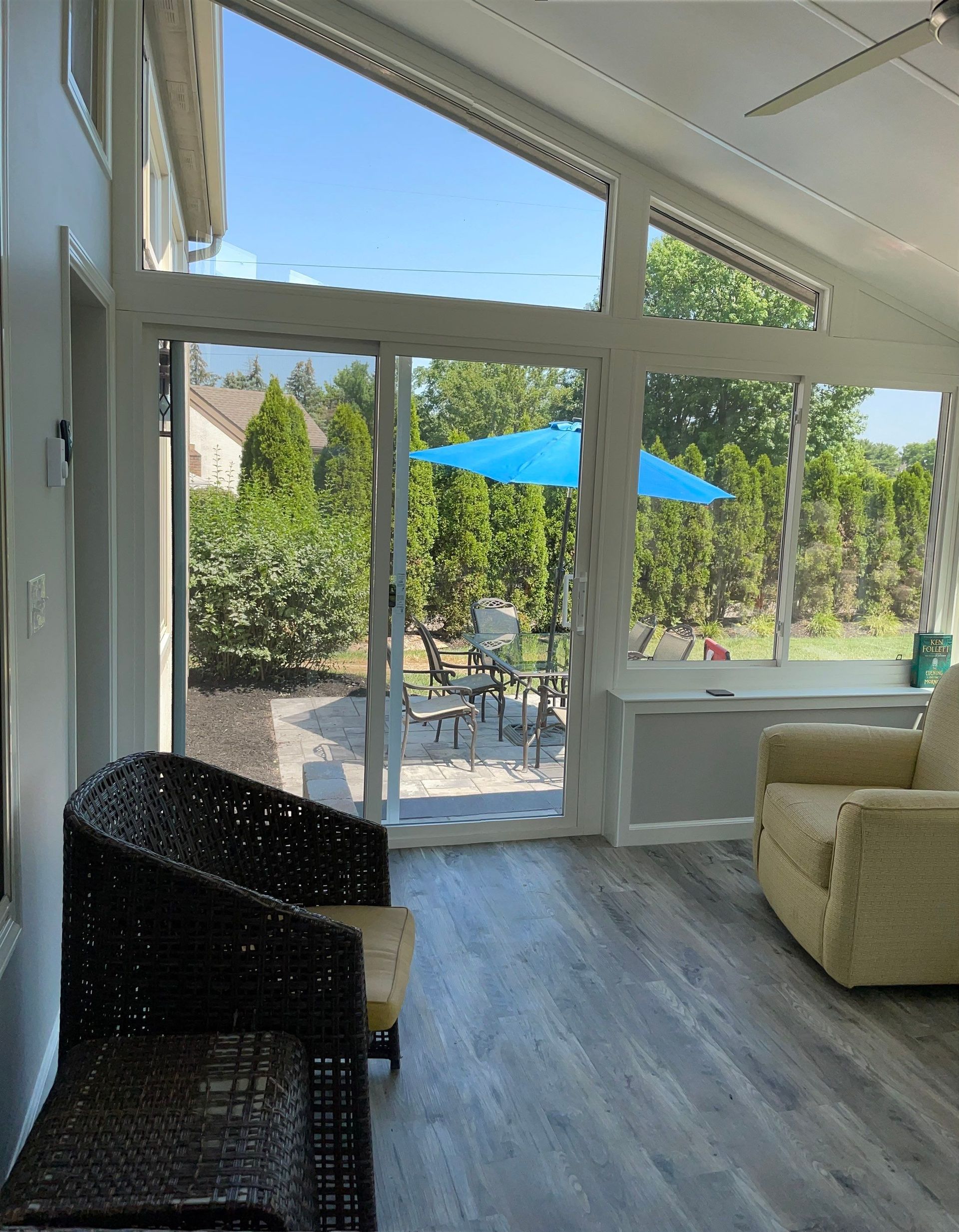 Bright sunroom with glass doors, wicker chair, beige sofa, and patio view with blue umbrella.