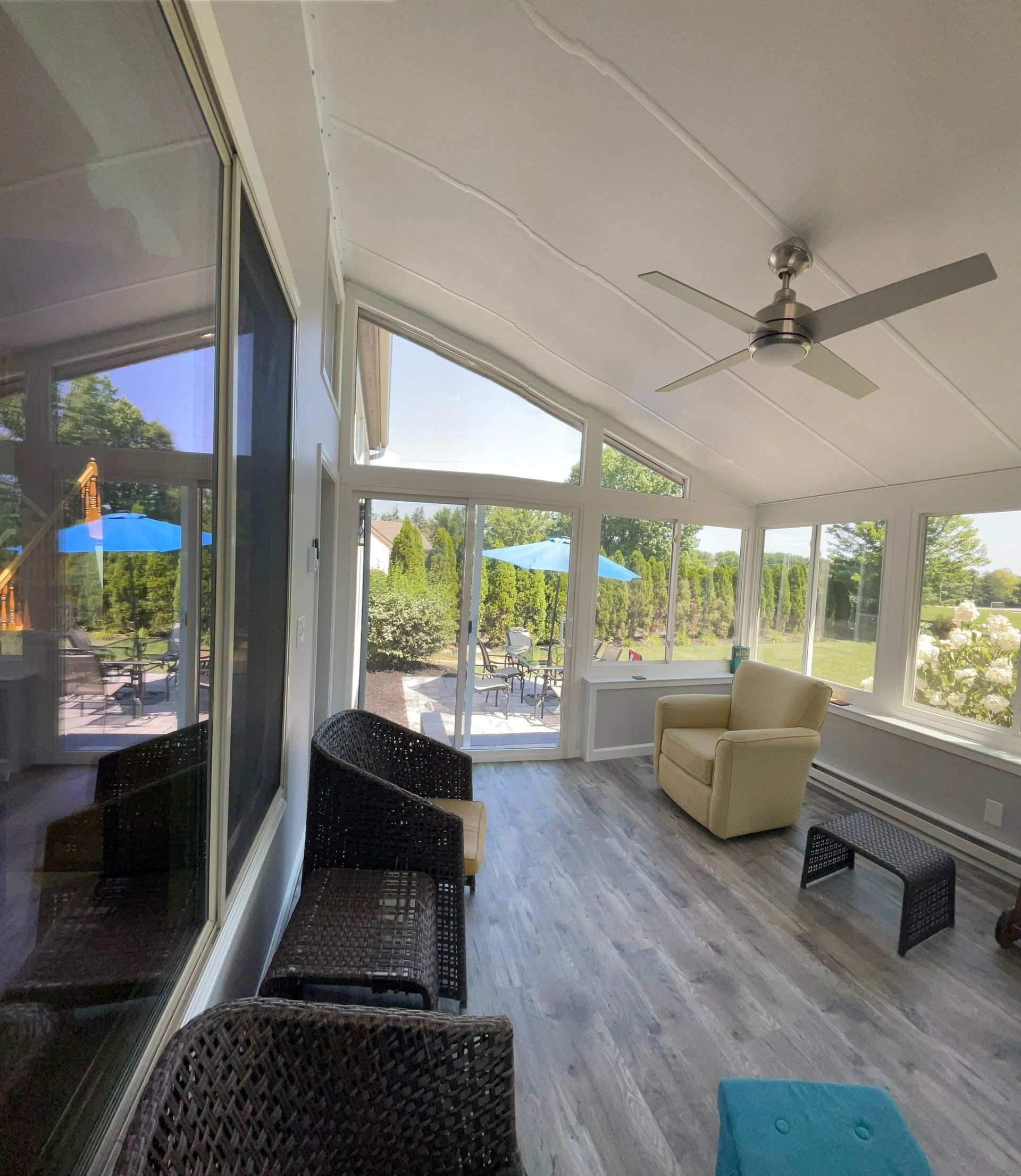 Sunroom with wicker seating, beige armchair, ceiling fan, and large windows overlooking trees and a patio