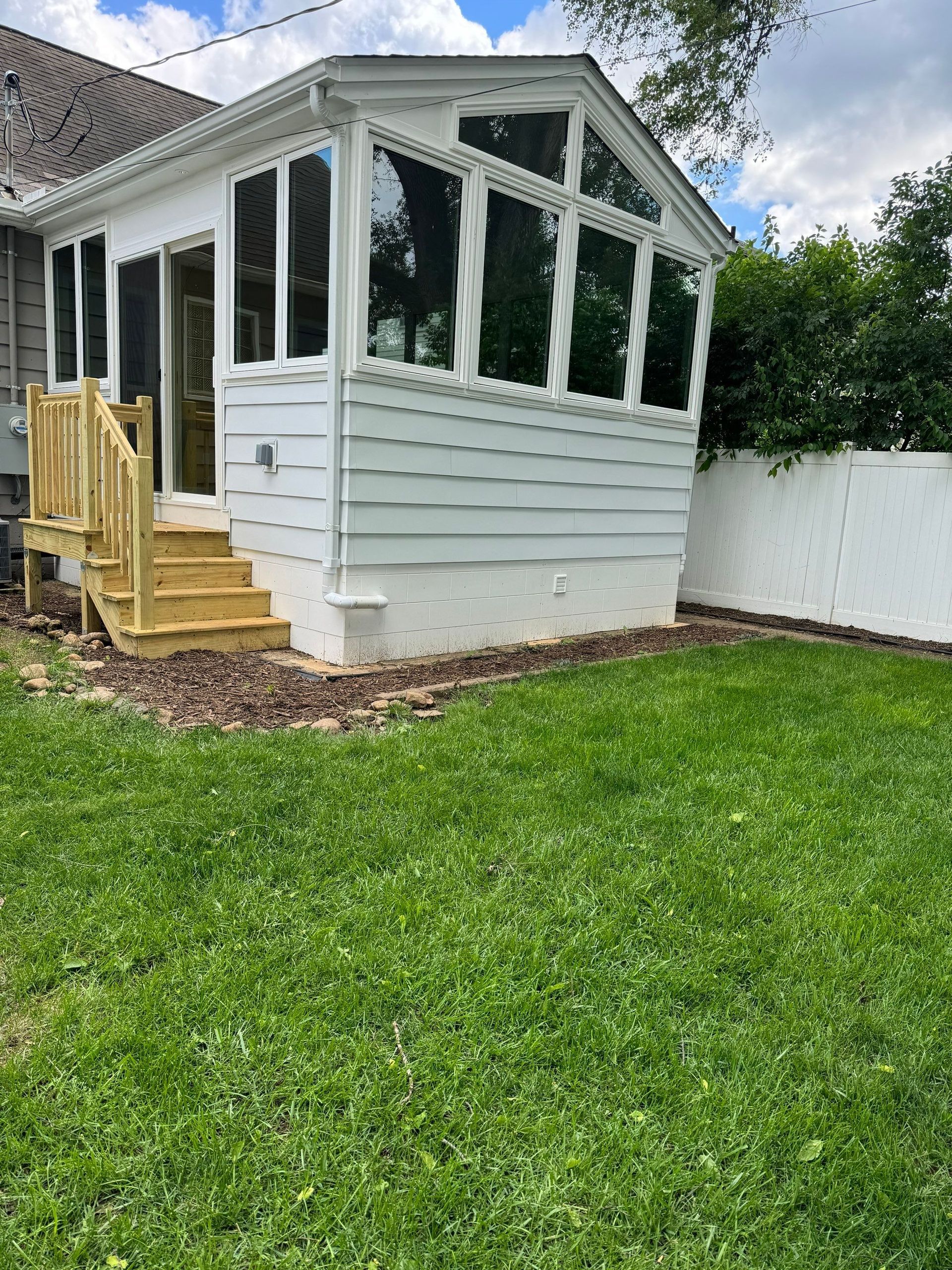 White sunroom addition with large windows, wooden steps, and a white fence in a grassy backyard.
