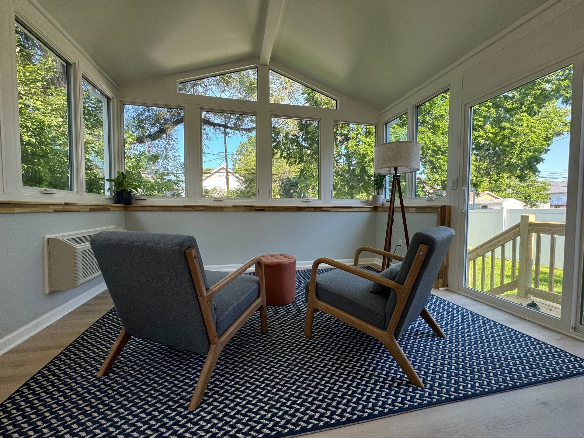 Covered sunroom with wicker chairs, patterned rug, and large windows overlooking a porch and trees.