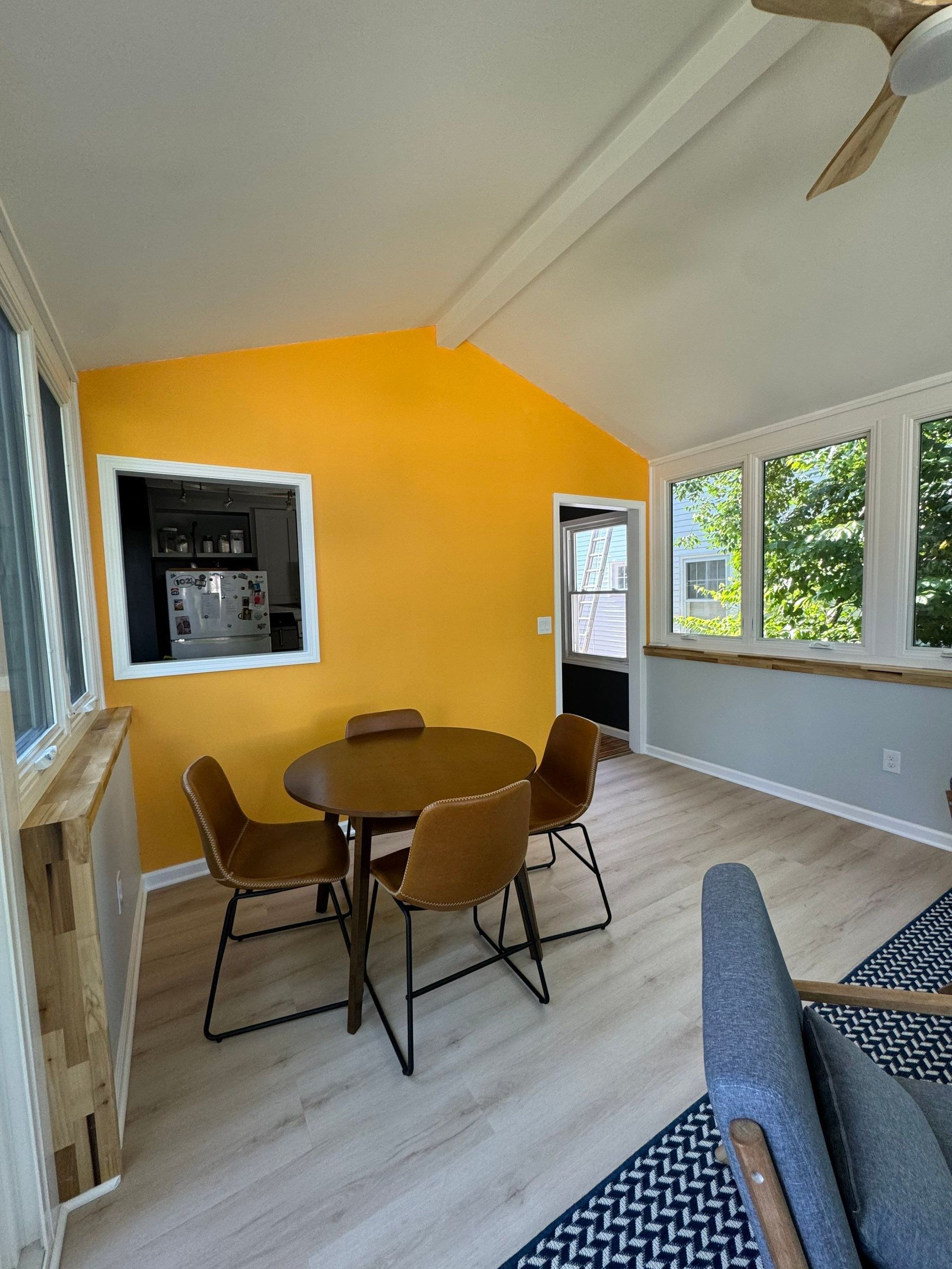 Bright sunroom with yellow accent wall, dining table and chairs, large windows, and gray sofa edge