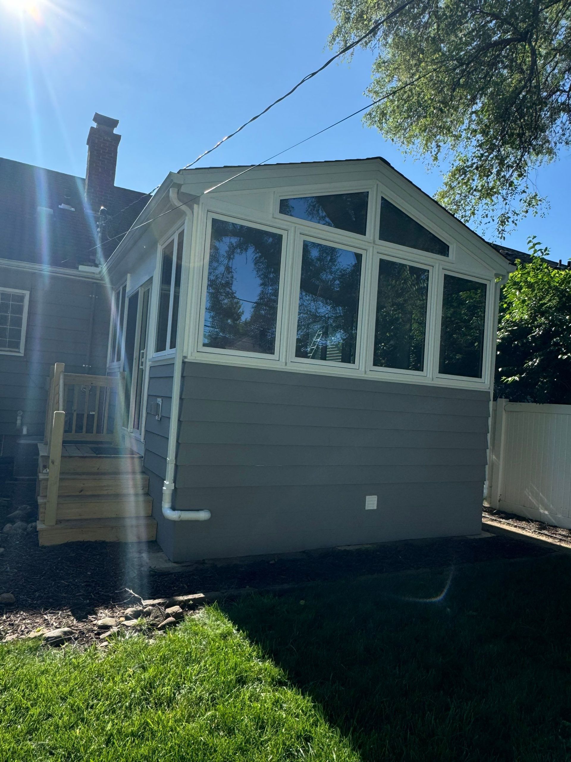 Gray sunroom addition with large windows on a house, beside a yard under a clear blue sky